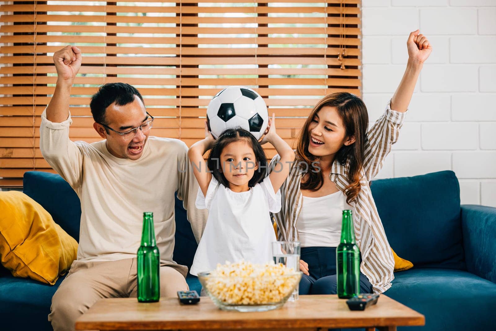 In the warmth of their home, parents and children watch a football match with popcorn and a ball, celebrating a goal. Their togetherness, smiles, and cheers reflect the joy of winning.