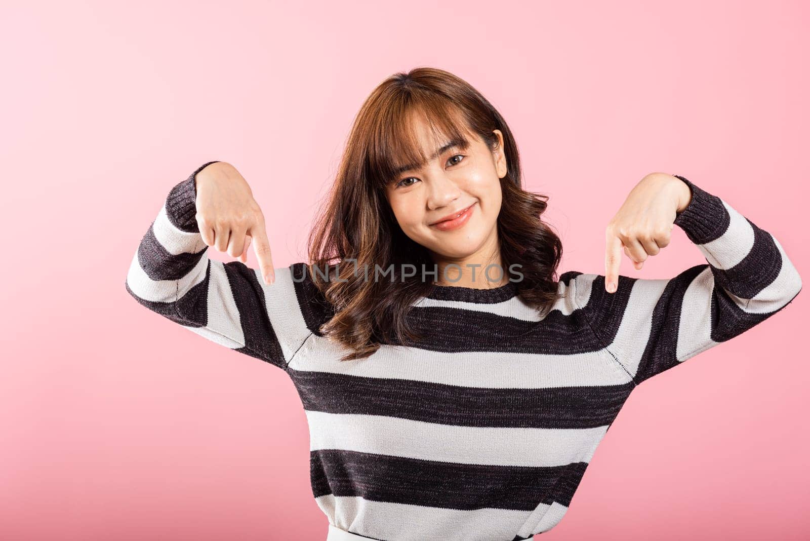 A happy Asian woman, in a portrait, smiles while making a two-finger pointing gesture downward. Studio shot isolated on pink with copy space, perfect for advertisements.