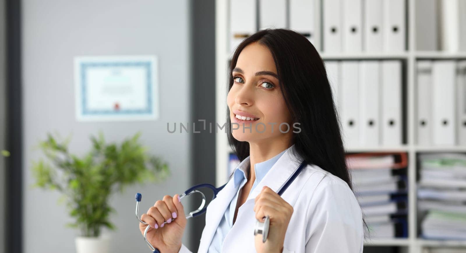 Beautiful smiling GP holding stethoscope posing in hospital office headshot