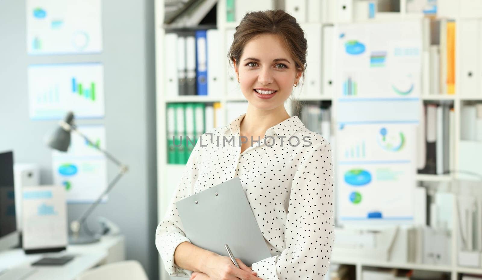Beautiful smiling woman standing in office holding document clipped to pad looking in camera headshot