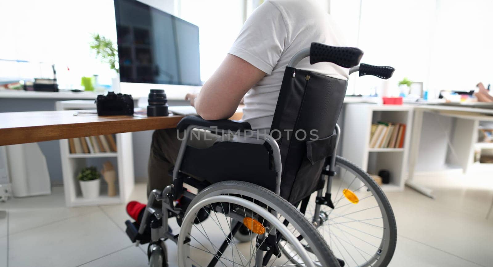 Close-up of man sitting in disabled carriage and looking in computer screen. Adapted workplace and office for people with disabilities. Camera on table. Fulfilling life. Professional specialist concept