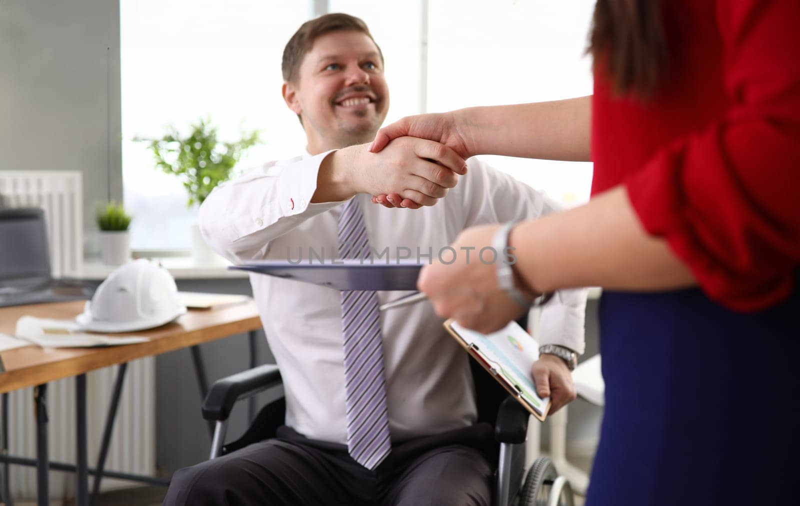 Close-up view of co-workers shaking hands and smiling to each other. Man in disabled carriage. Working process at company office. Employee holding folder with papers. Business concept