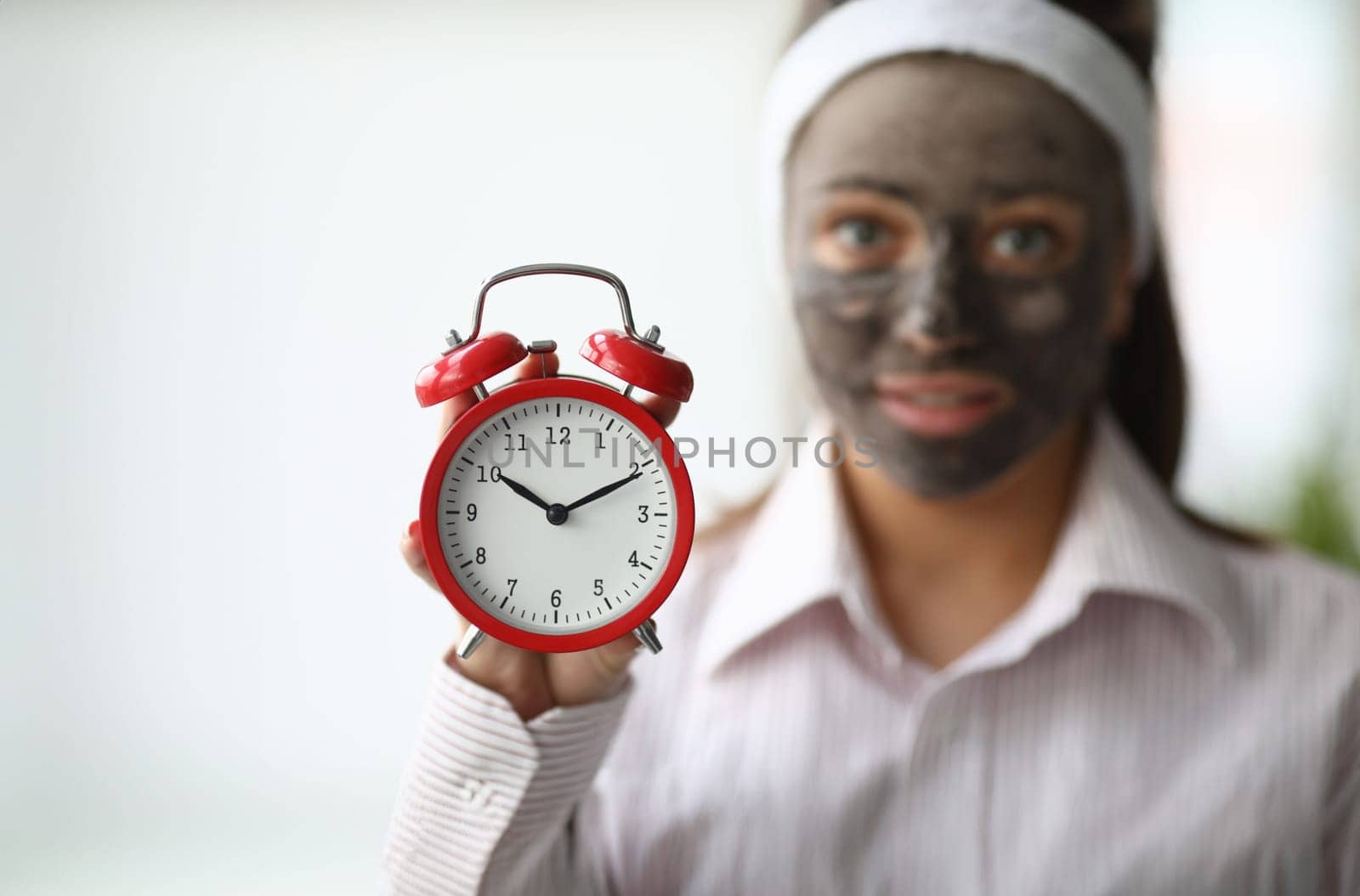 Close-up view of woman holding red clock. Young female put timer to take off moisturizing clay mask. Spa day at home. Wellness and beauty skincare treatment concept