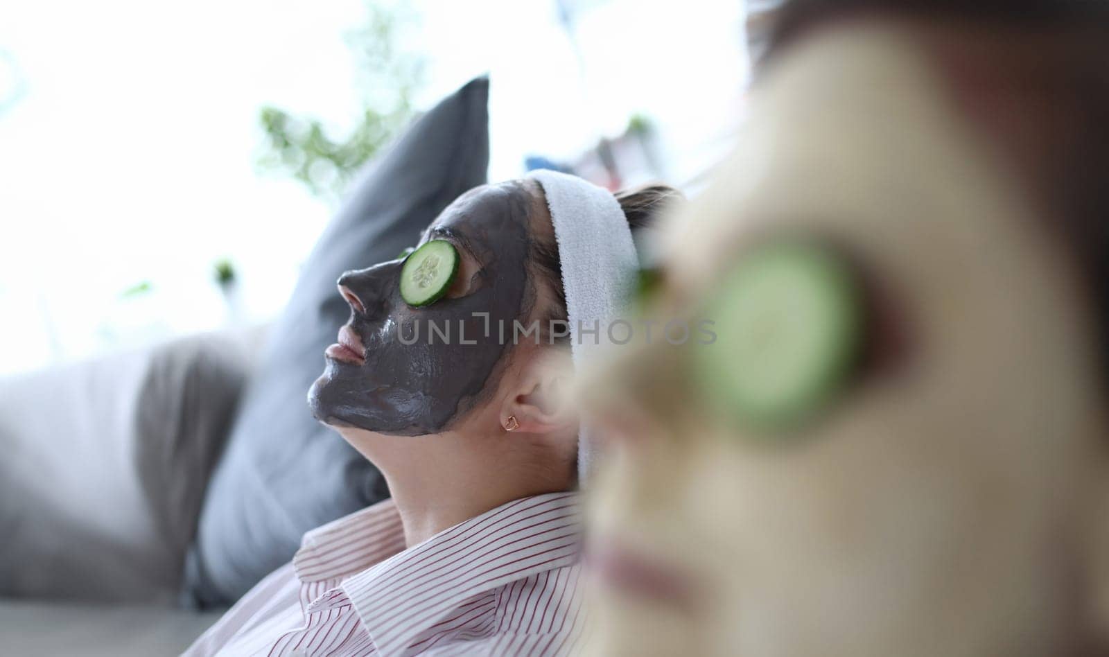 Close-up view of young woman relaxing on sofa with eyes closed wearing clay face mask. Girl and friend having spa day. Skincare treatment and beauty concept