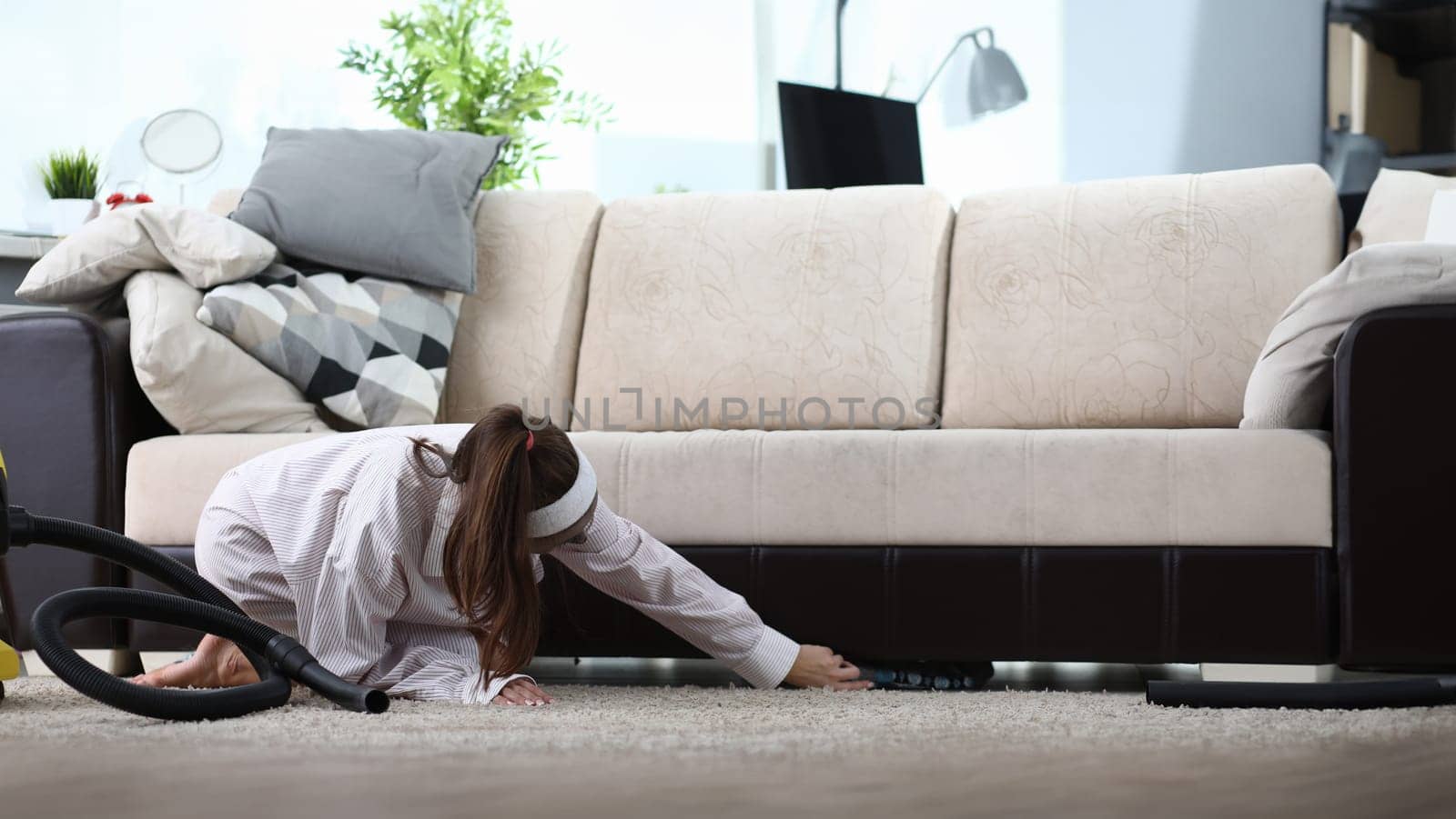 Close-up of young woman cleaning in living room. Female in leisure wear find cloth under sofa. Vacuum cleaner and pillows on couch. Big cleanup day concept