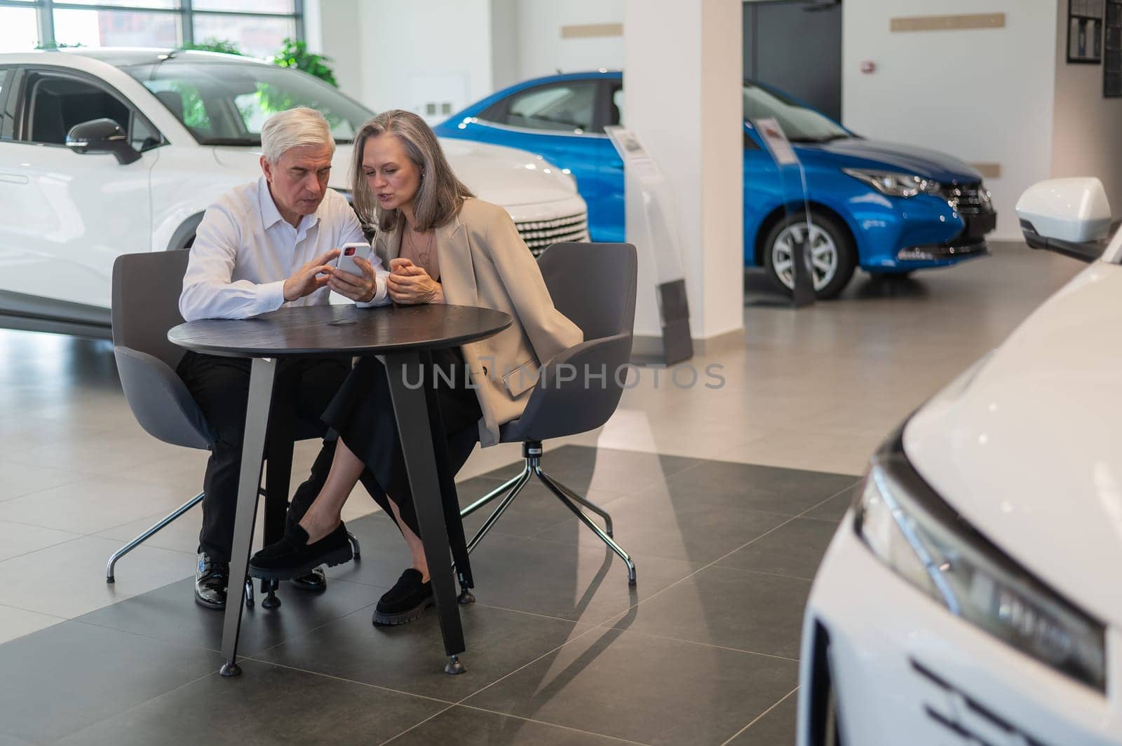 A mature couple is sitting in a car dealership and looking at a smartphone. by mrwed54
