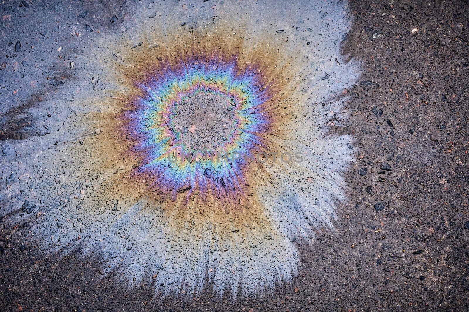 Close-up of an iridescent oil or gasoline spill on a wet asphalt, viewed from above. Bold multicolored spots on the asphalt