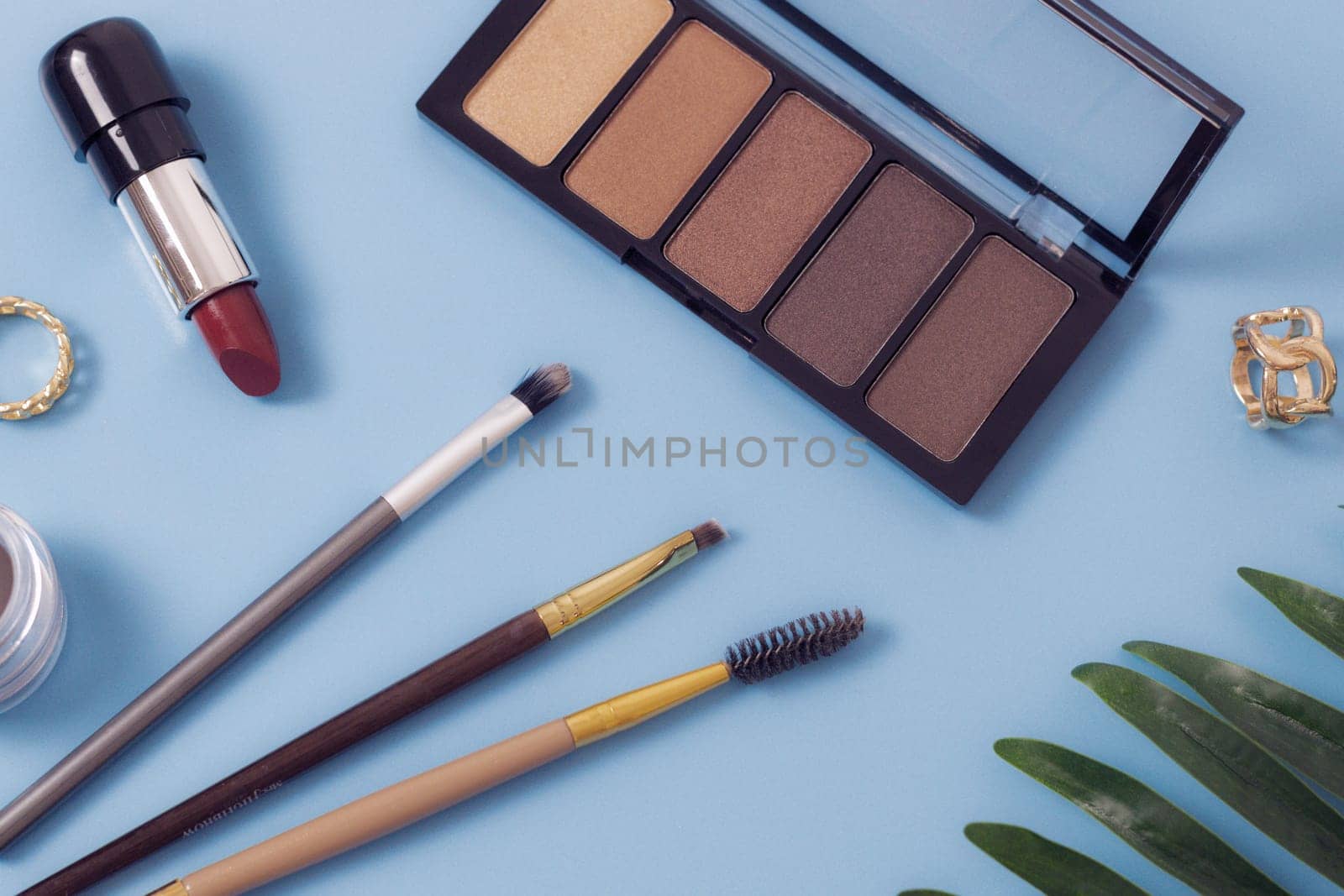 A set of cosmetics from a palette of nude shades of eye shadow, red lipstick, makeup brushes, soft eyebrow shadows and two gold rings with a palm branch on a blue background, flat lay close-up. The concept of cosmetics, beauty salon.