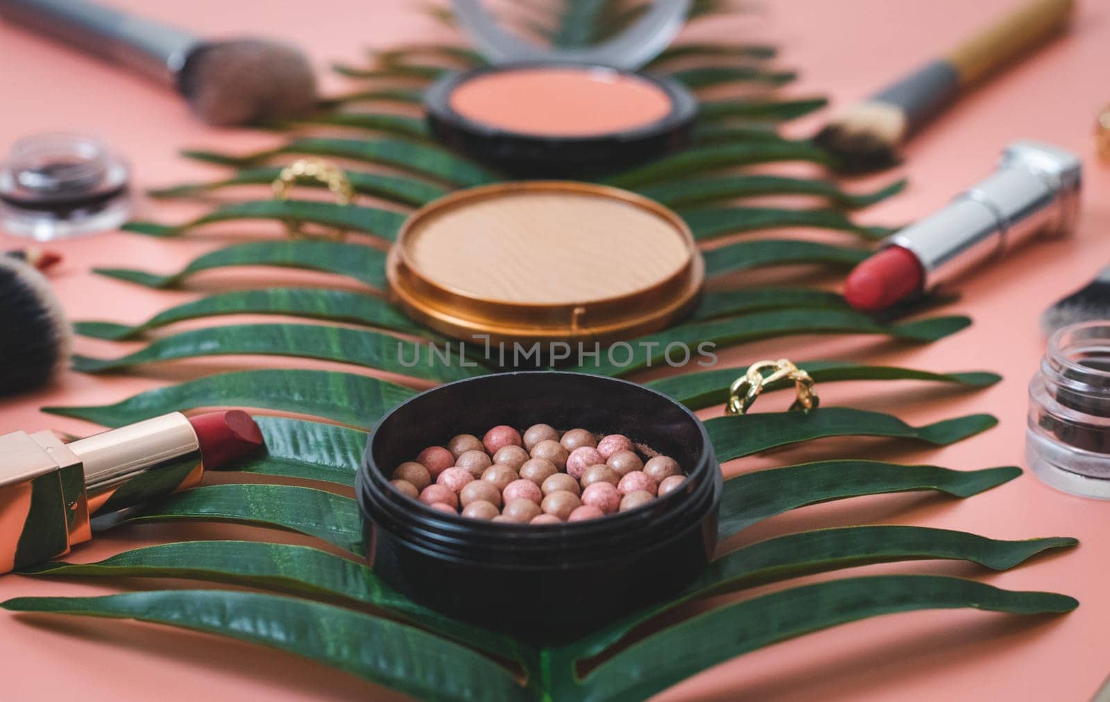 Granular face powder box, gold ring, red lipsticks, makeup brushes and hard texture powder boxes in blur on a palm branch on a pink background, side view close-up of the field. The concept of cosmetics, beauty salon.