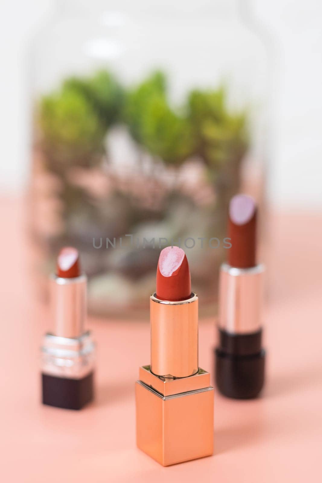 Three pieces of red lipstick stands on a pink and white background with a blur of the decoration of a plant in a jar, side view close-up with depth of field. The concept of cosmetics, beauty salon.