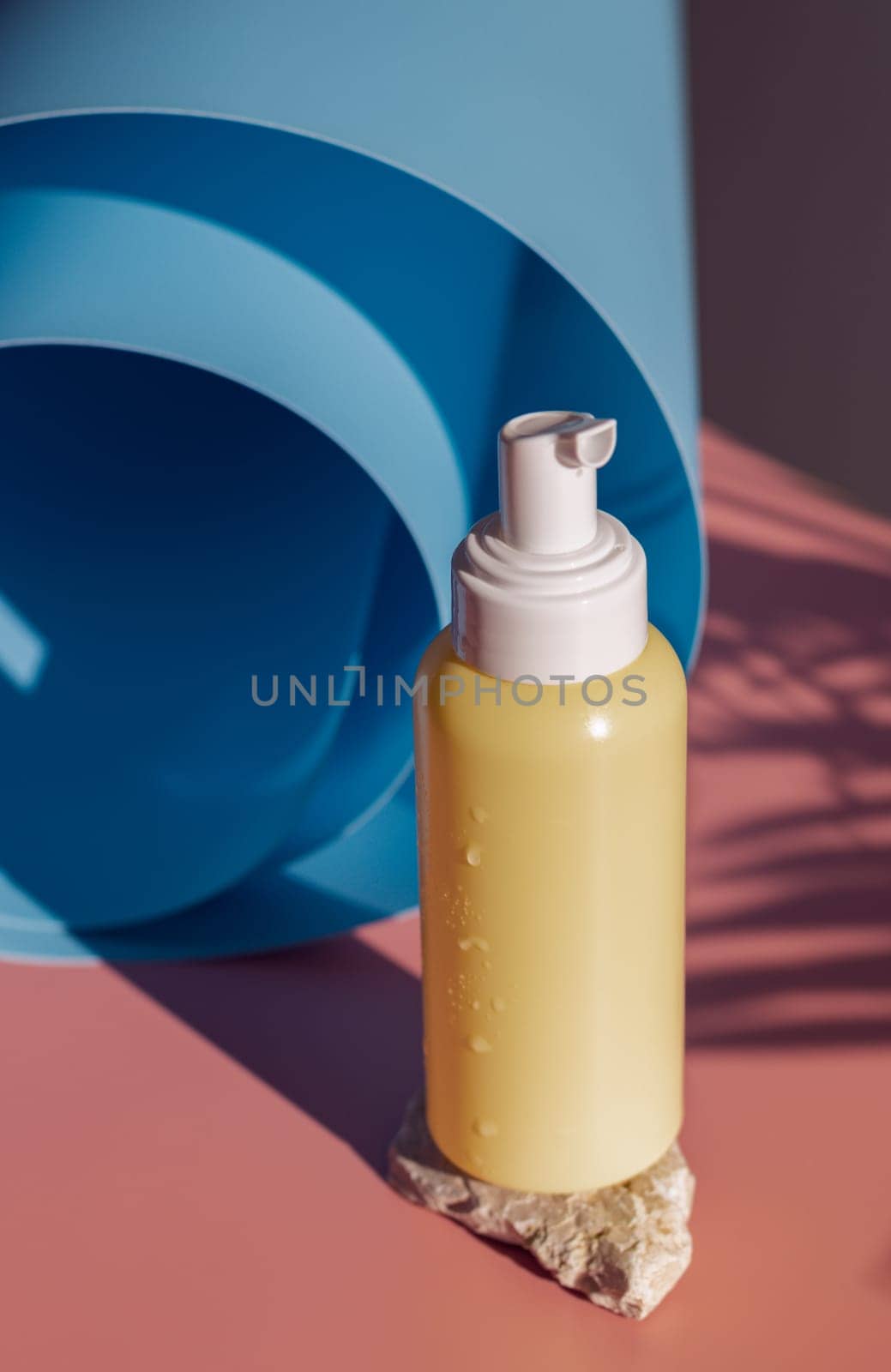 One small yellow bottle with a dispenser, drops of water stands on a stone near a twisted blue roll on a pink background with a palm tree branch, close-up top view. Cosmetics concept, body care.