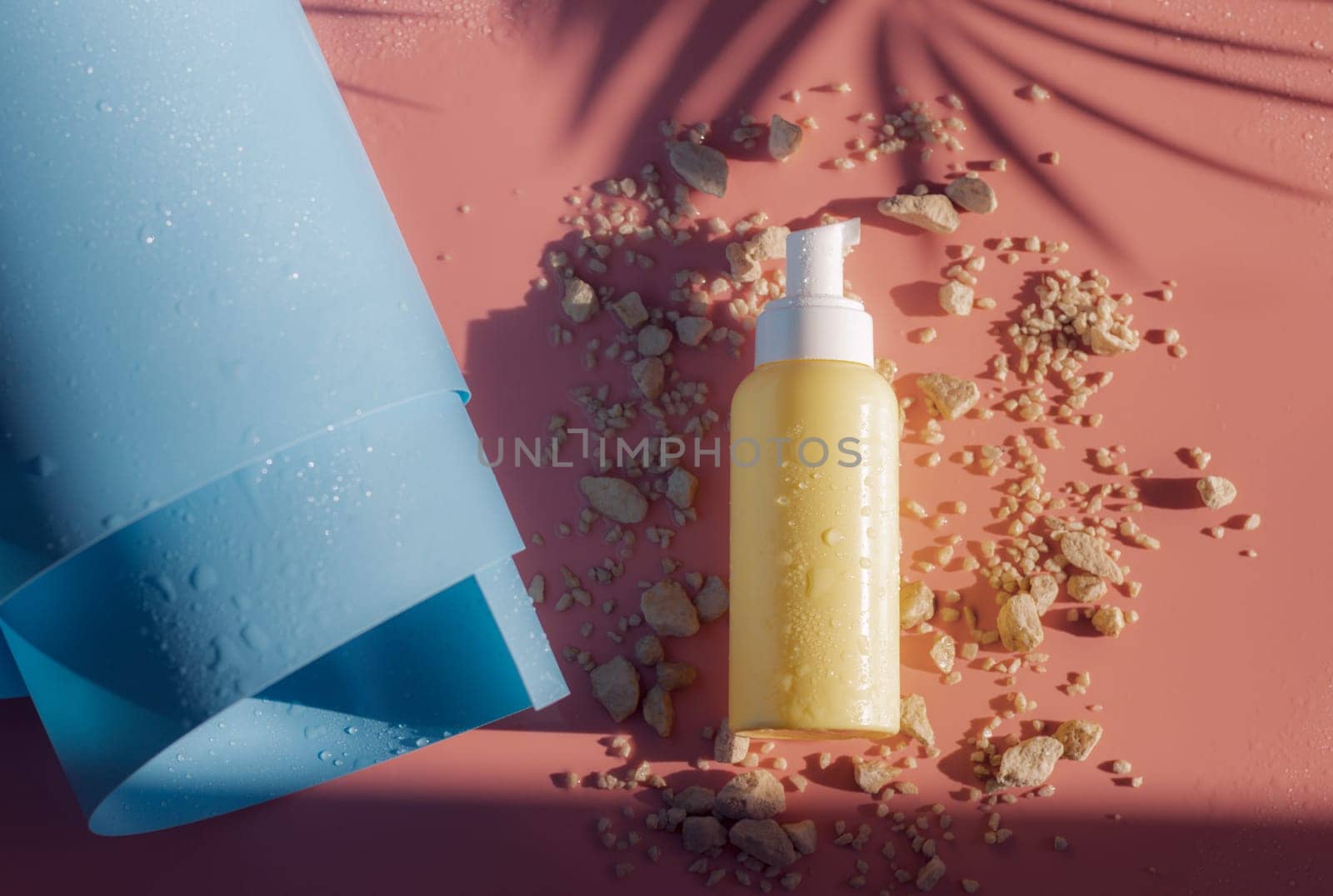 One small yellow bottle with a dispenser, drops of water lies on small stones near a twisted blue roll on a pink background with a shadow of a palm tree branch, flat lay close-up. Cosmetics concept, body care.