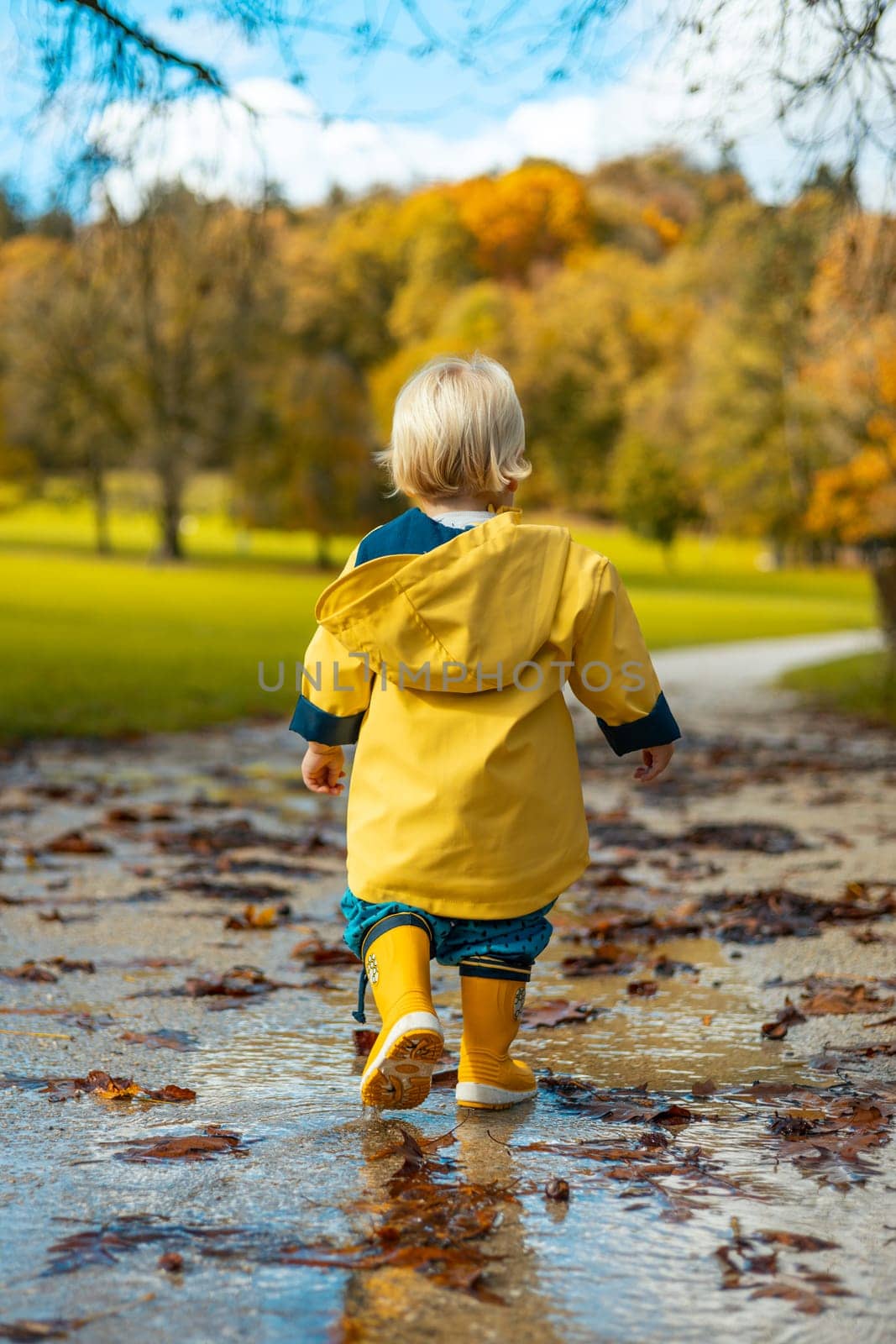 Sun always shines after the rain. Small bond infant boy wearing yellow rubber boots and yellow waterproof raincoat walking in puddles in city park on sunny rainy day. by kasto