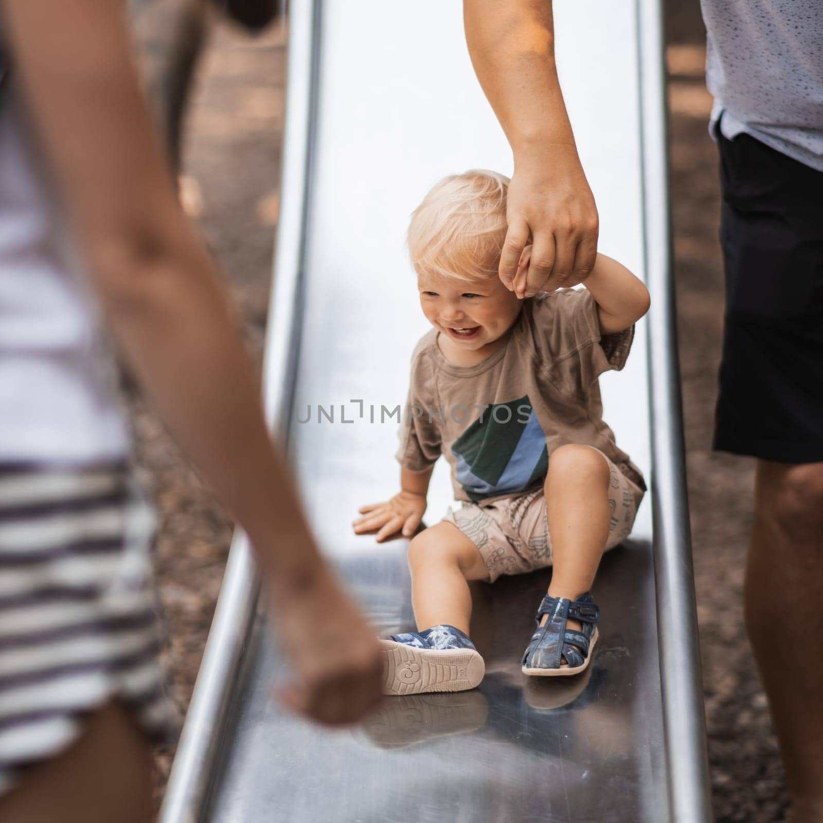 Parent hand holding little infant baby boy child while sliding on urban playground on a sunny summer day. Family joy and happiness concept.