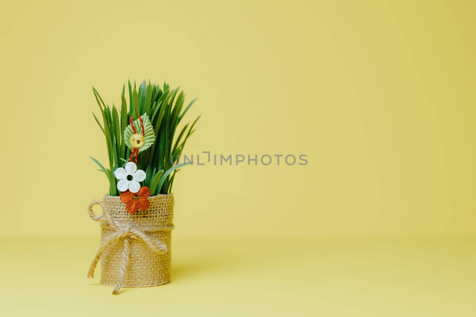 One beautiful homemade martisor of two flowers, a petal and a cheerful smiley face hanging on sprouted wheat in a jute pot stands on the left on a pastel yellow background with copy space on the right, side view close-up.