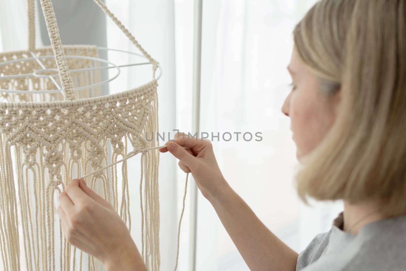 Woman making macrame lampshade using white cord to tie the strings together. Woman knits lampshade for interior using macrame technique. Closeup of woman knits boho chandelier at home