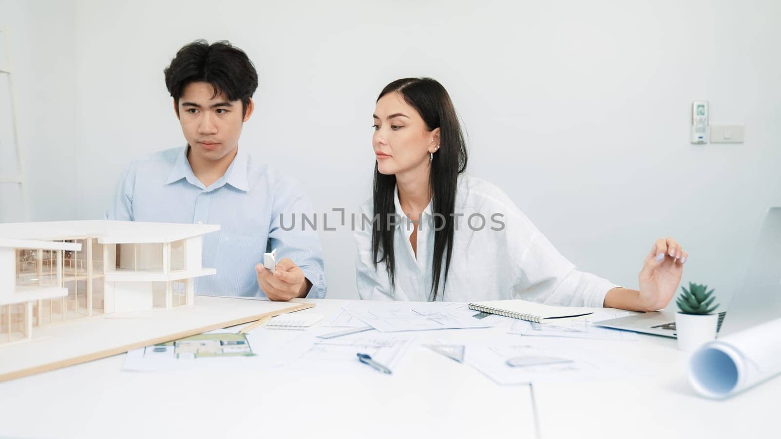 Professional male engineer measure house model by using ruler while beautiful cooperative coworker working together by using laptop to analysis data. Creative design and teamwork. Closeup. Immaculate.