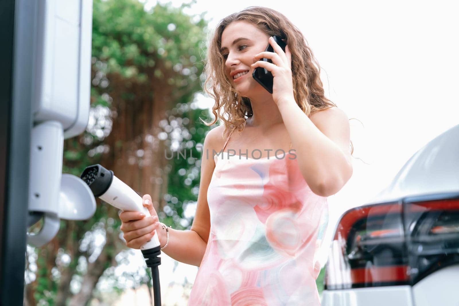 Eco-friendly conscious woman recharging modern electric vehicle from EV charging station. EV car technology utilized as alternative transportation for future sustainability. Synchronos