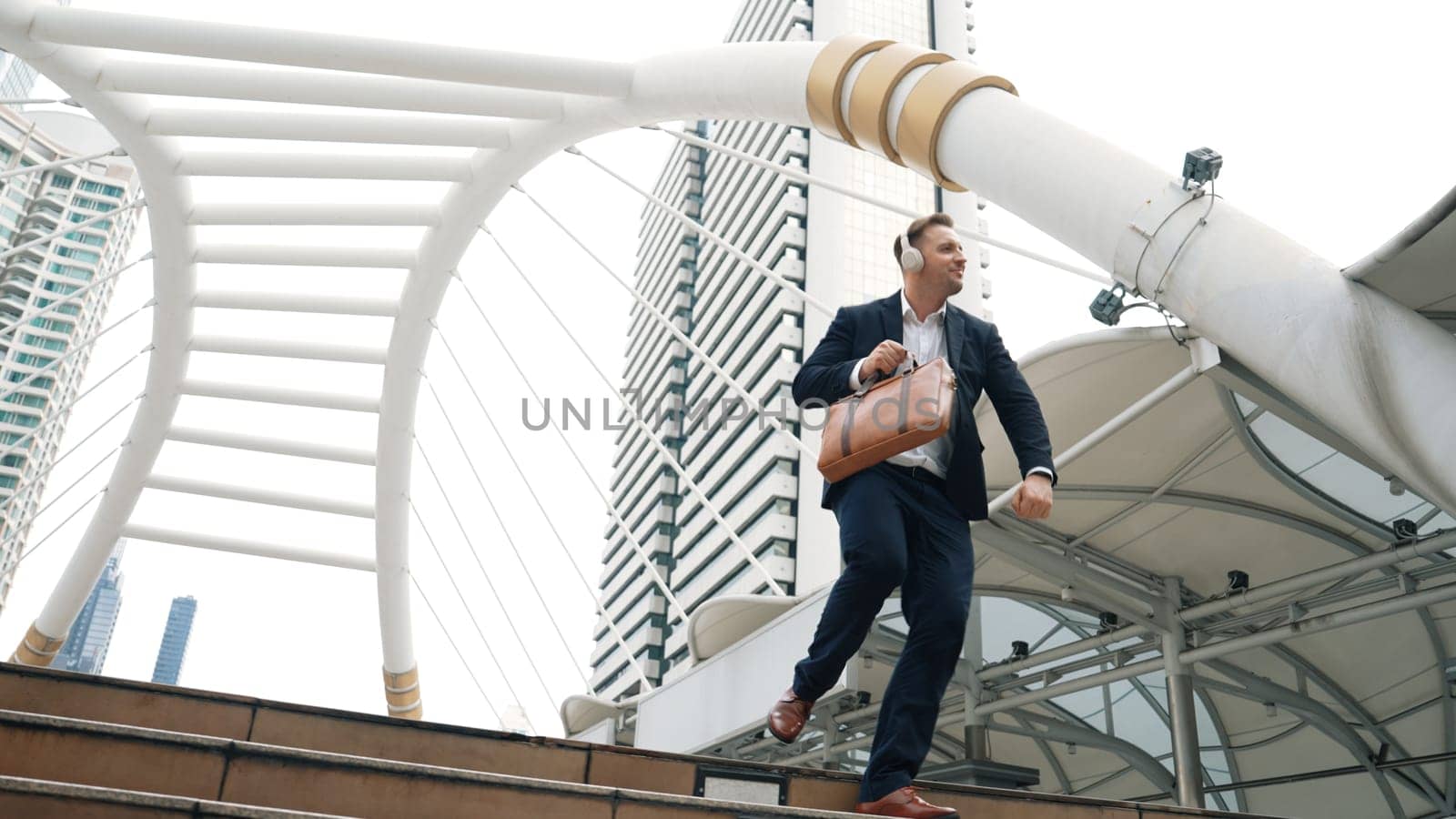 Happy businessman walking down stair and moving to music from headphone. Low angle camera view of smart manager listen song by using heaset and going back to home in urban city in lively mood. Urbane.