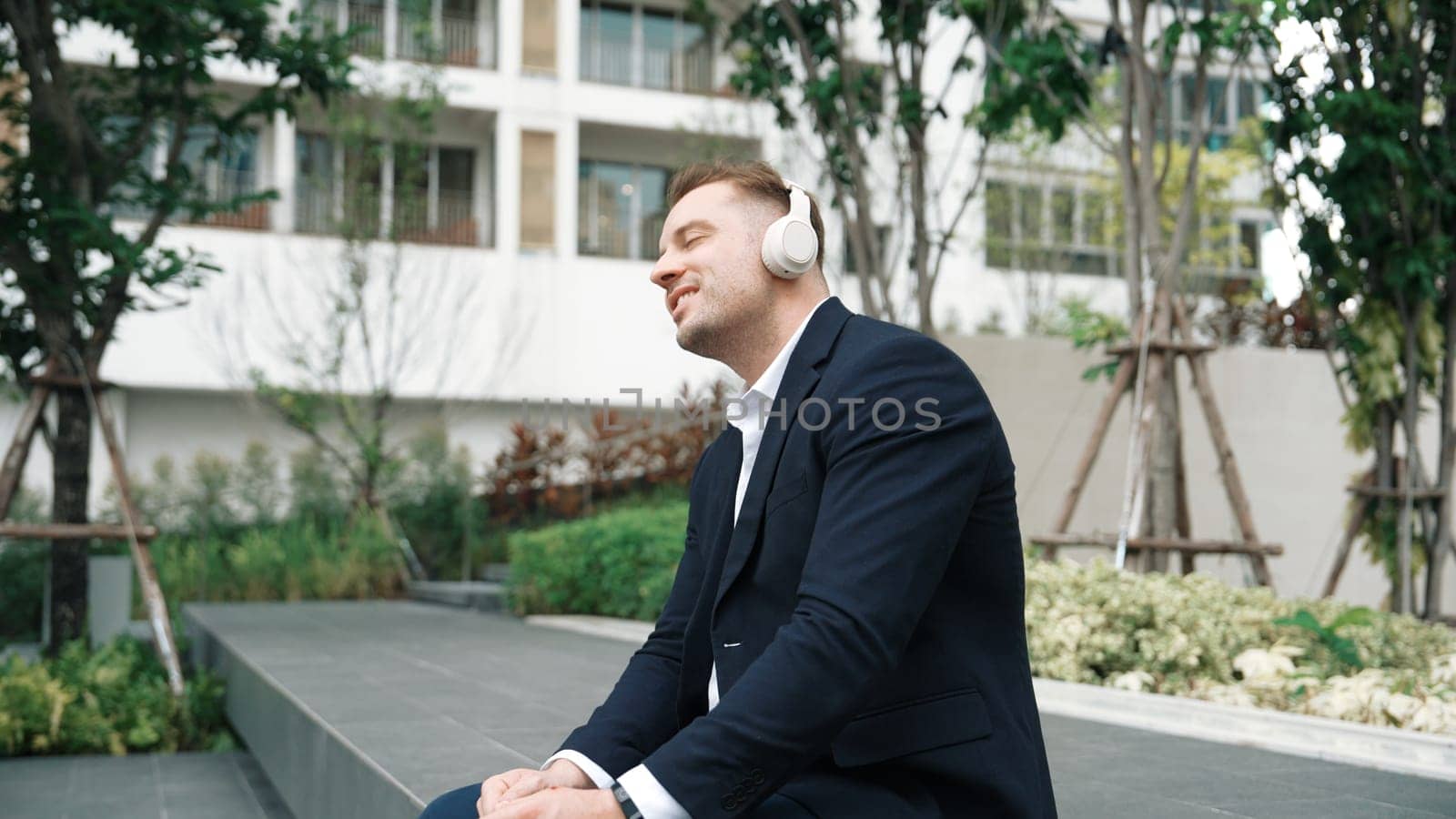 Professional businessman wearing headphone and while moving to relaxing music in green city. Caucasian project manager dancing to song while sitting and relaxed at eco urban city. Lifestyle. Urbane.