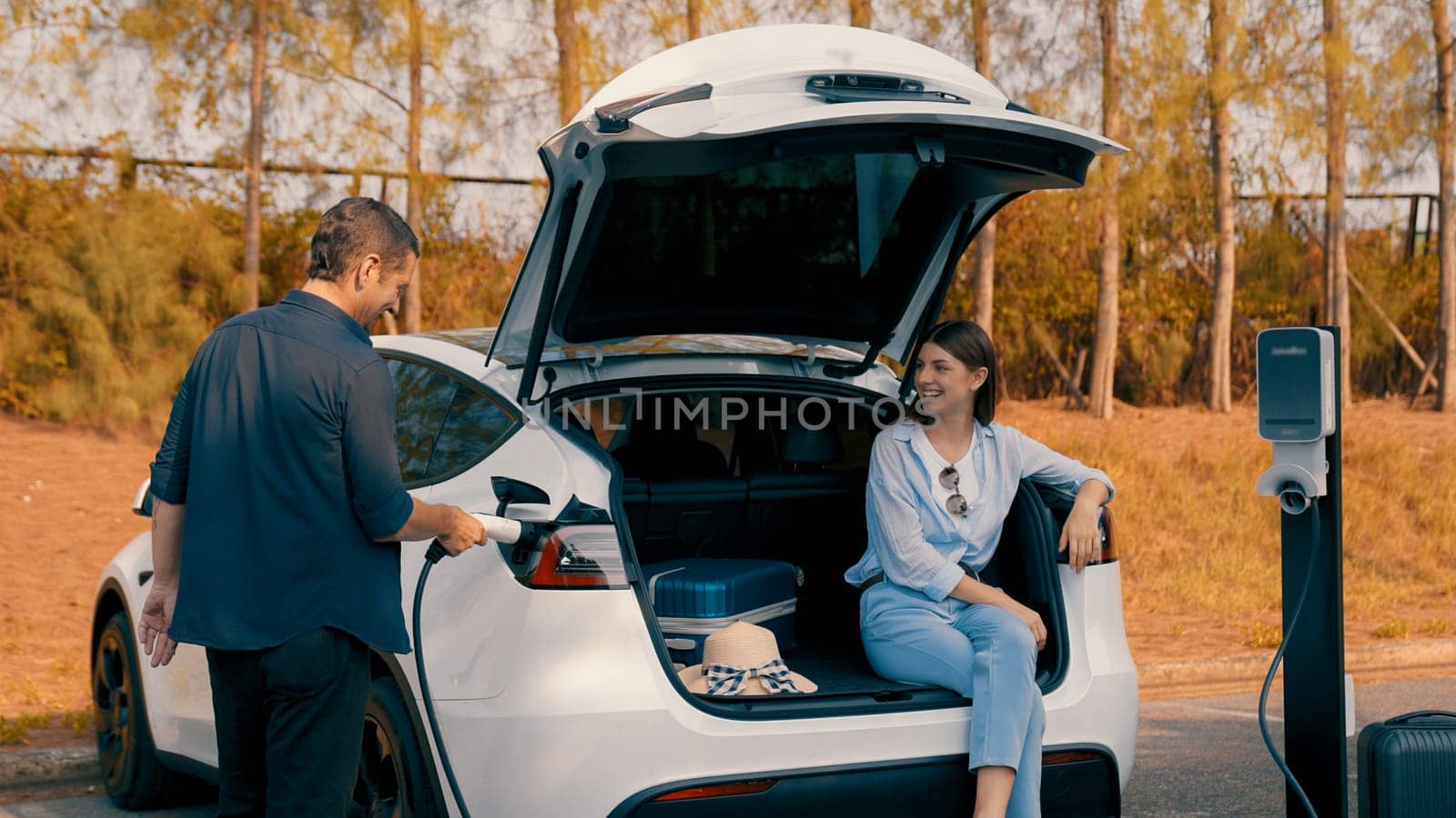 Couple recharge their EV car with electric battery charging station at rest stop during their road trip travel during autumnal season to national park. Relaxing holiday with eco friendly car. Exalt