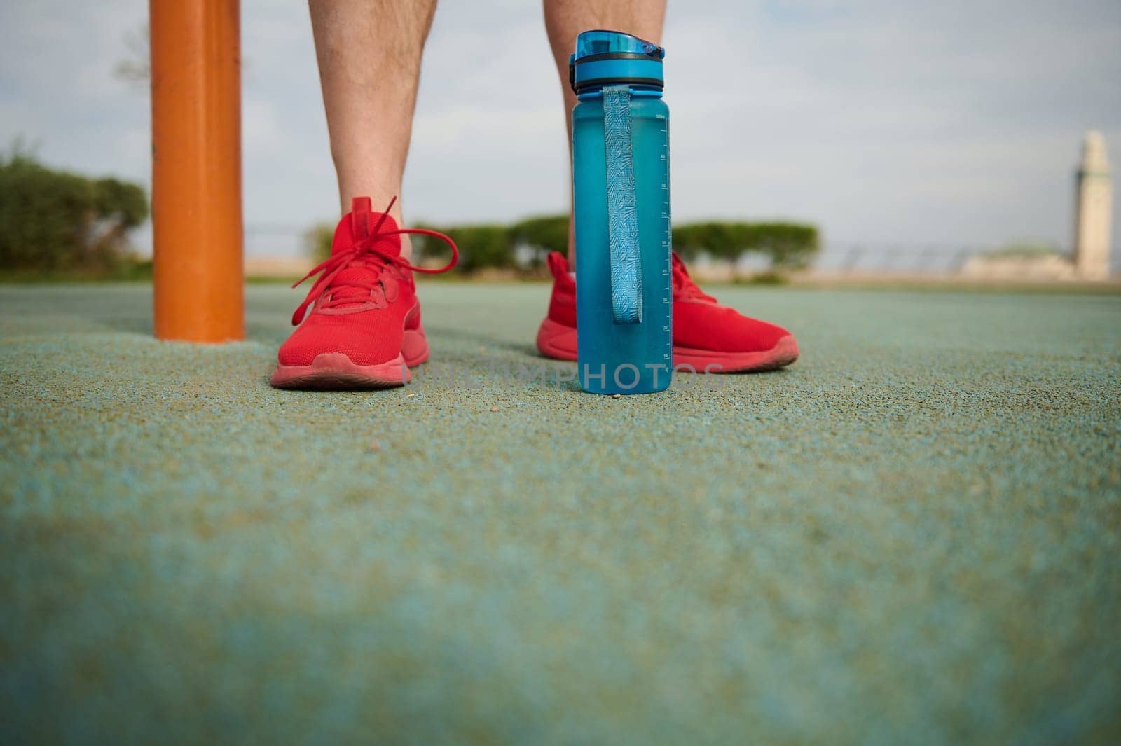 Cropped view of athlete's feet in red sneakers and bottle of water on the sportsground. Copy ad space by artgf