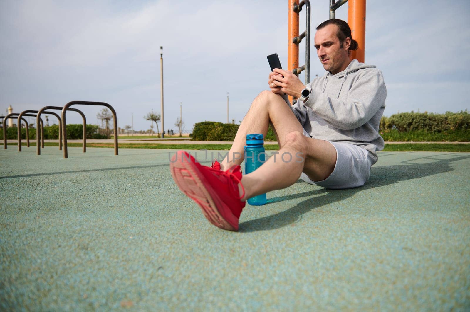 Athletic man checking mobile sports app on his smartphone, analyzing his workout results while sitting on the sportsground after a bodyweight training outdoor. People. Sport and modern technologies