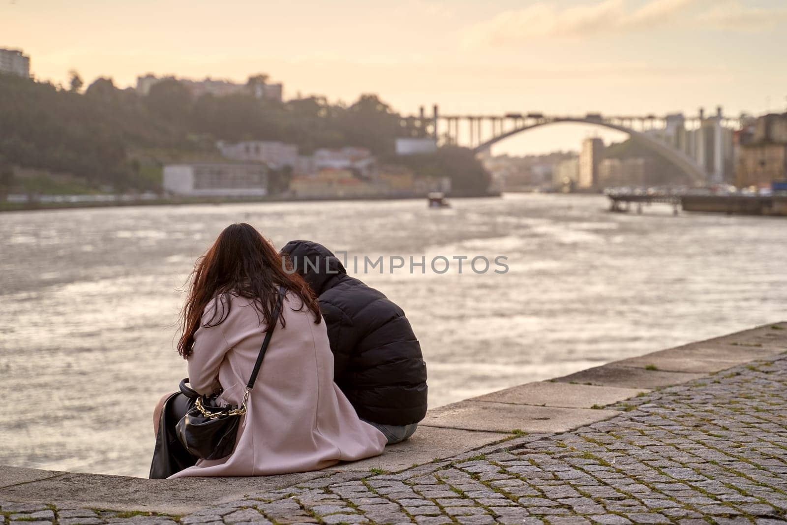 Porto, Portugal: Februray 22 2024 - Young couple sitting on a bank of the Douro river with Arrabida Bridge on background