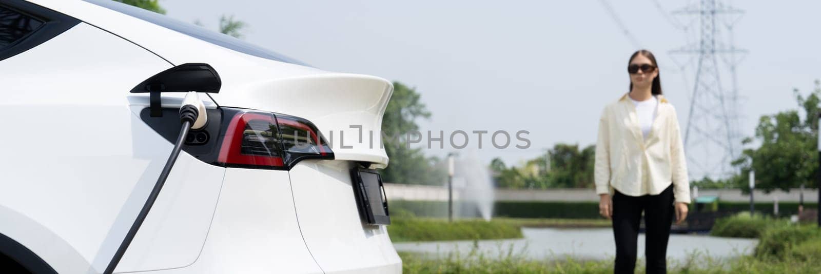 Young woman recharge EV car battery at charging station connected to power grid tower electrical industrial facility as electrical industry for eco friendly vehicle utilization. Panorama Expedient