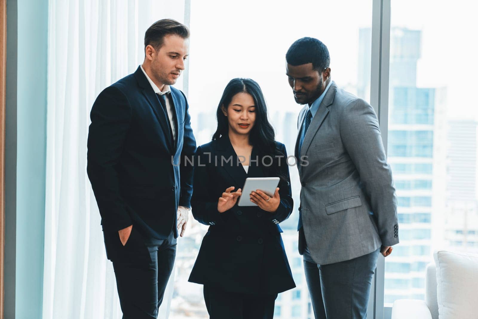 Diverse smart businesspeople standing near window with skyscraper while looking at business data analysis. Group of business team using tablet represent marketing plan, strategy, idea. Ornamented..