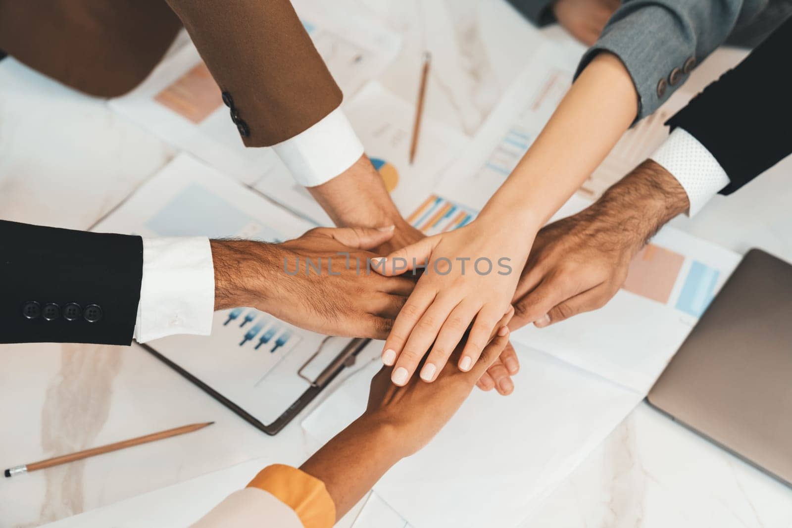 Top view of corporate diverse businesspeople putting hands together on meeting table with document scatter around at business meeting room. Represented unity, cooperation, collaboration. Ornamented.