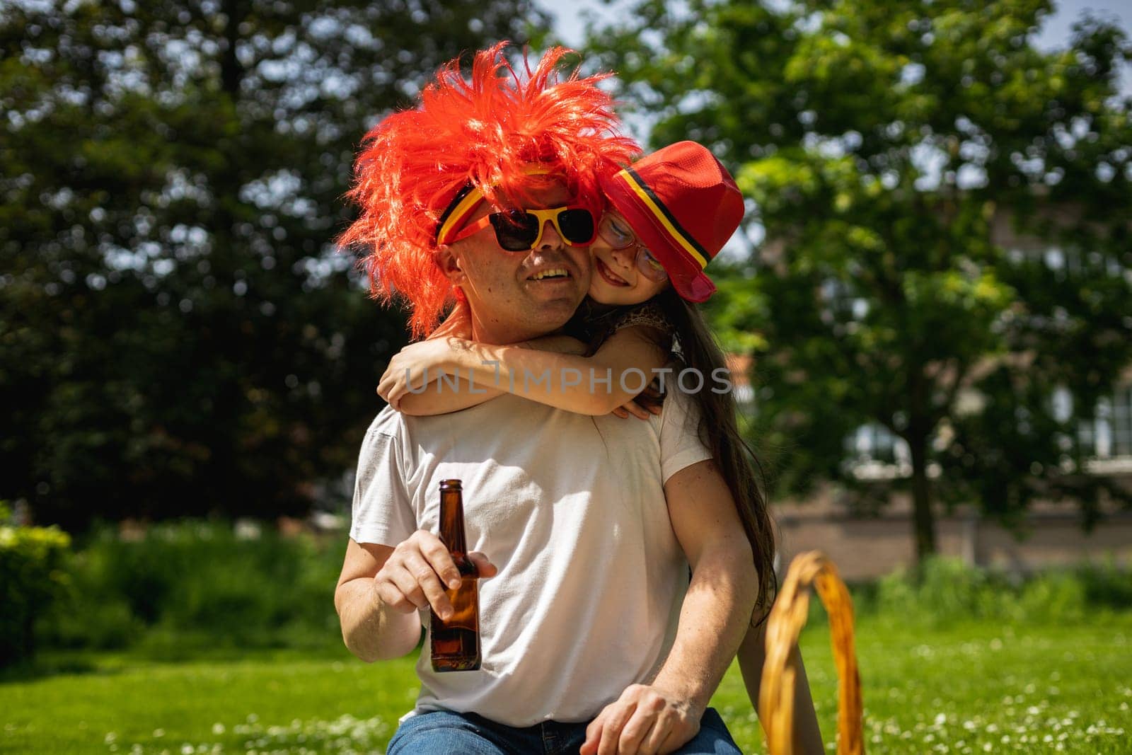 Portrait of handsome young caucasian man in sunglasses with belgian flag, red wig holding bottle of beer in hands and happy daughter hugging his neck, sitting on lawn and celebrating belgium day in city park on sunny summer day, close up side view.