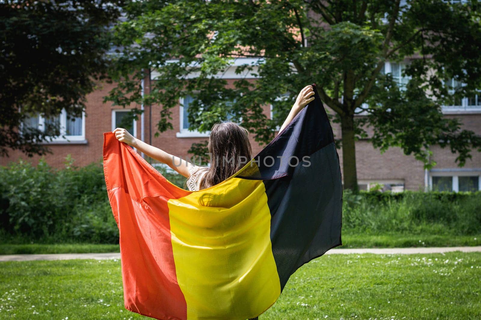 Portrait of one beautiful Caucasian little brunette girl with loose hair standing from the back and holding the Belgian flag open in a city park on a summer day, close-up side view.