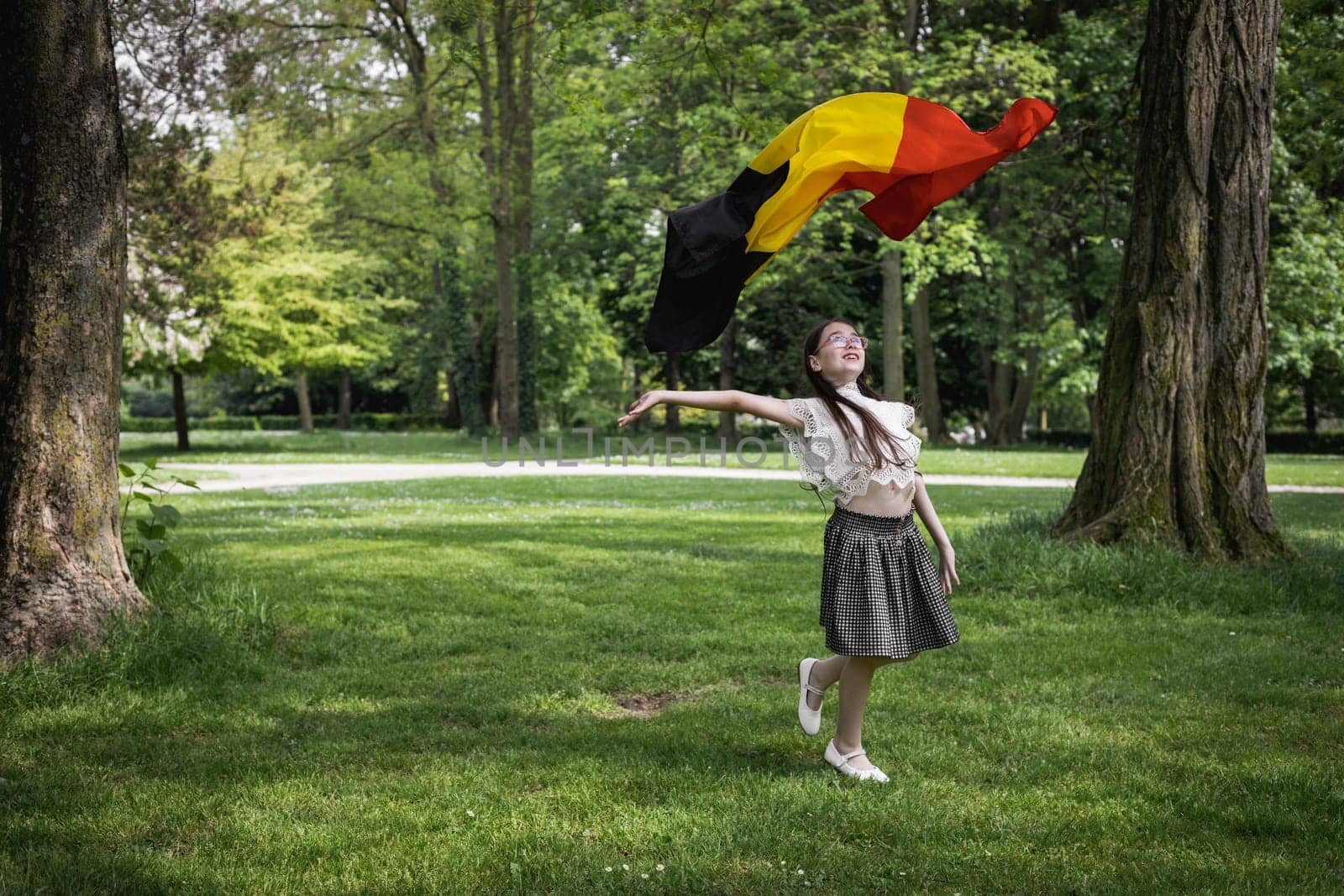 Portrait of one beautiful Caucasian little brunette girl in a skirt with her hair down, having fun, throwing up the Belgian flag in a city park on a summer day, close-up side view with copy space on the left.