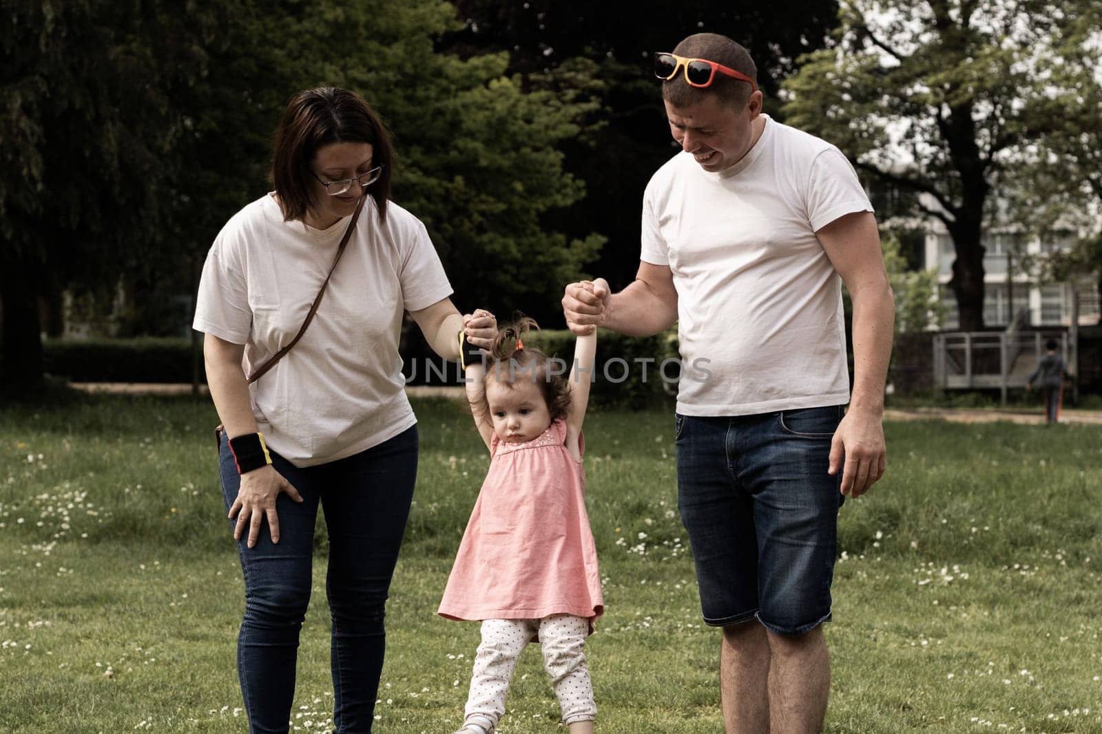 Portrait of one beautiful Caucasian young parents raising their little daughter by two hands in a pink dress with a ponytail on the top of her head while standing in a park on a summer day, side view close-up.
