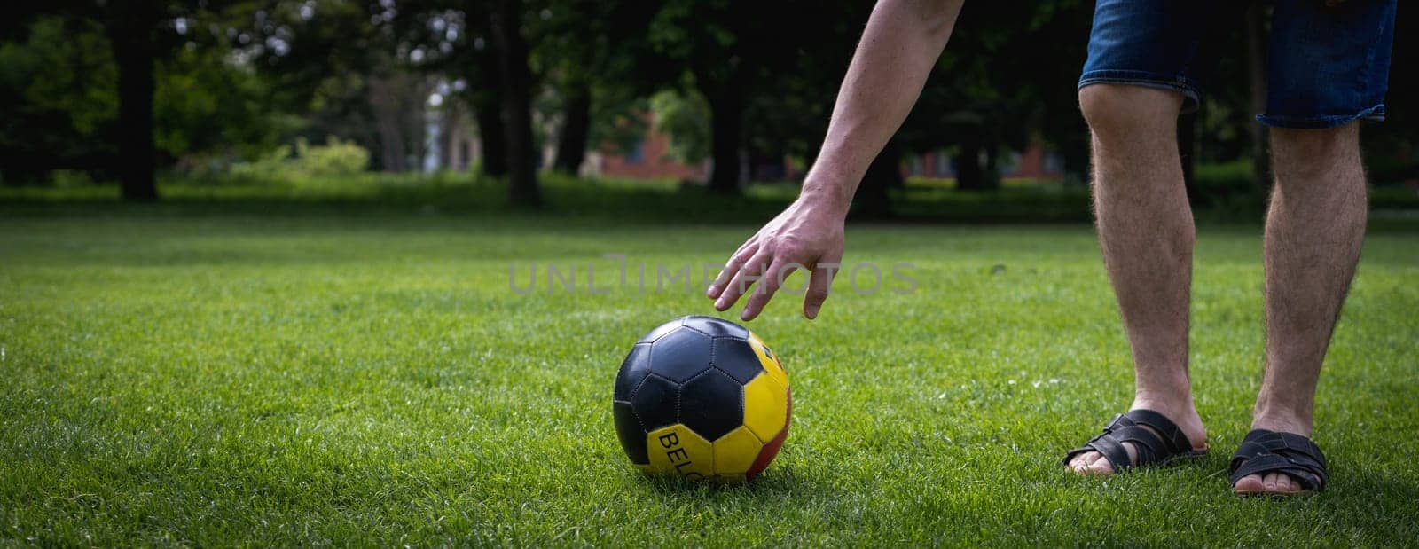 An unrecognizable young man Caucasian man reaches for a soccer ball with Belgium written on it, lying on the green lawn of a city park on a summer day, close-up view from the side.