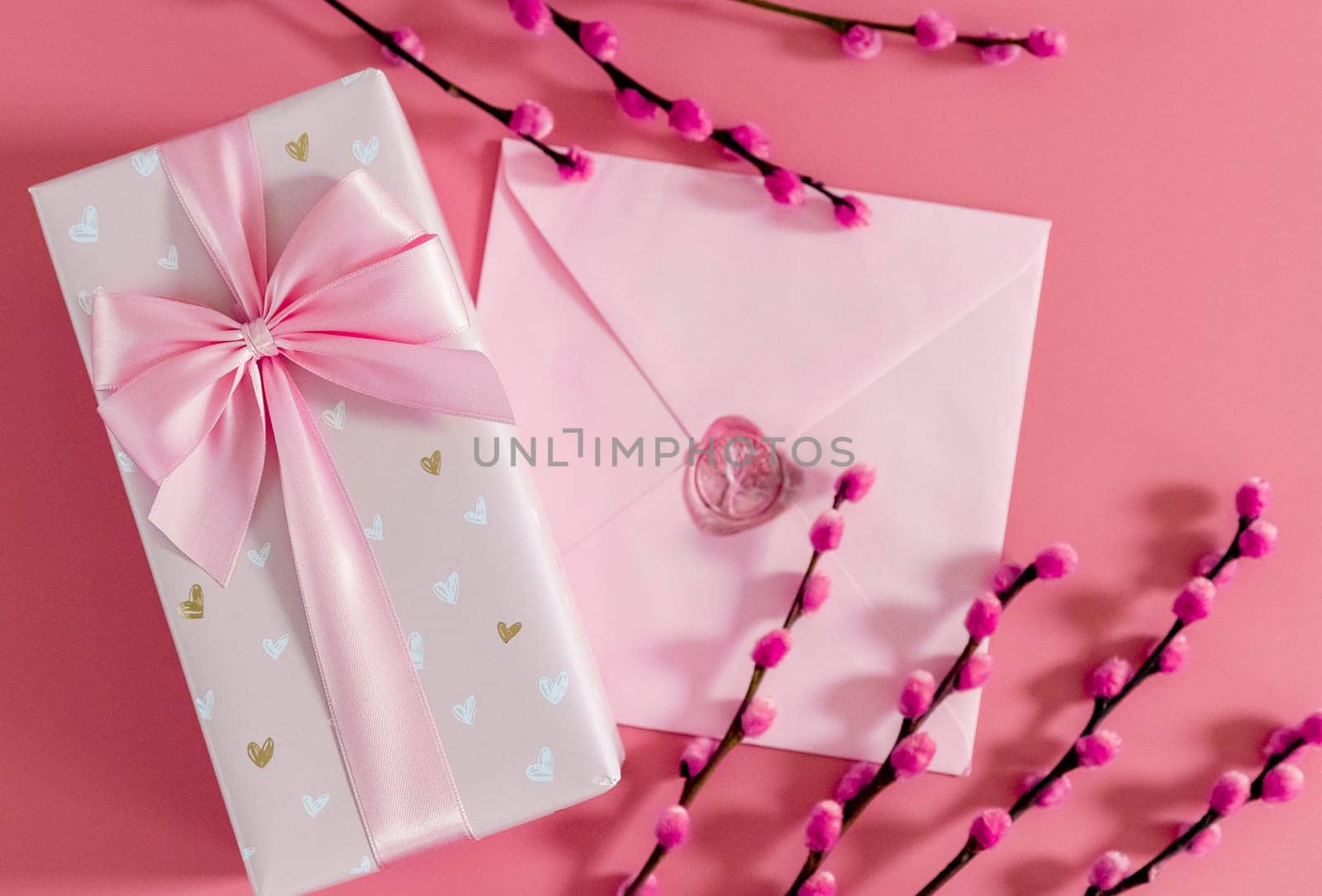 One beautiful gift box with a bow, a sealed envelope and willow branches lie on a pink background, flat lay close-up with depth of field.