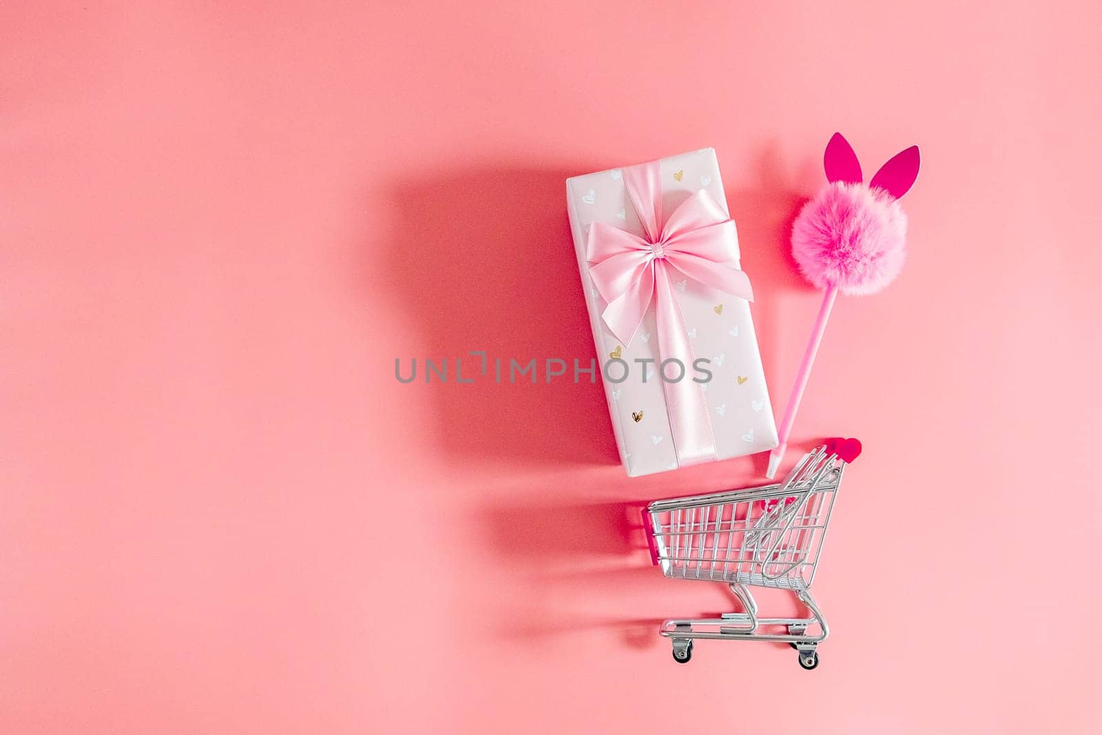 One large beautiful gift box with a bow and a fluffy bunny handle falling out of a mini shopping cart on the right on a pink background with copy space on the left, flat lay close-up.