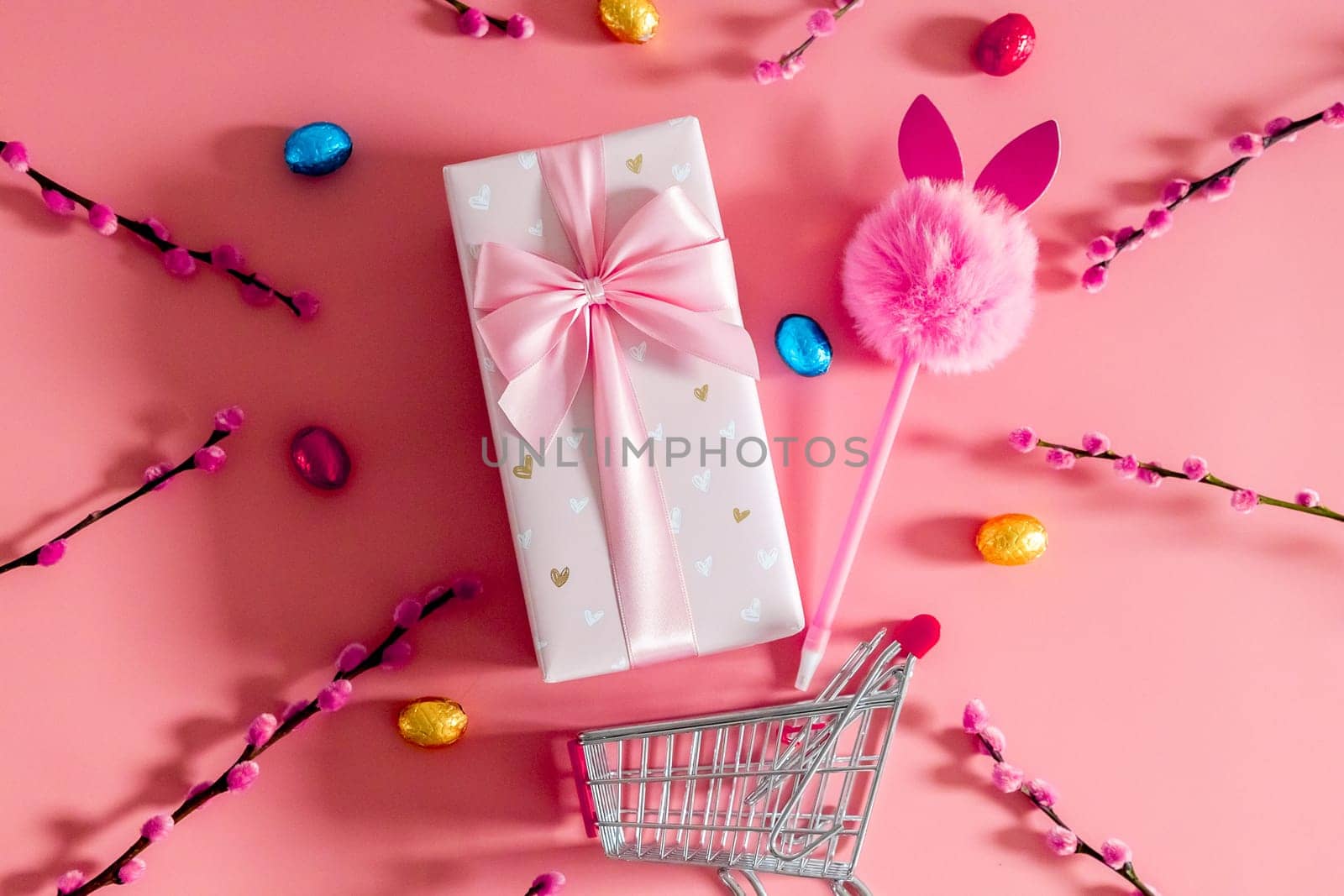 One large beautiful gift box with a bow, a fluffy bunny handle, chocolate Easter eggs in multi-colored wrappers and willow branches fall out of a mini shopping cart on a pink background, flat lay close-up.