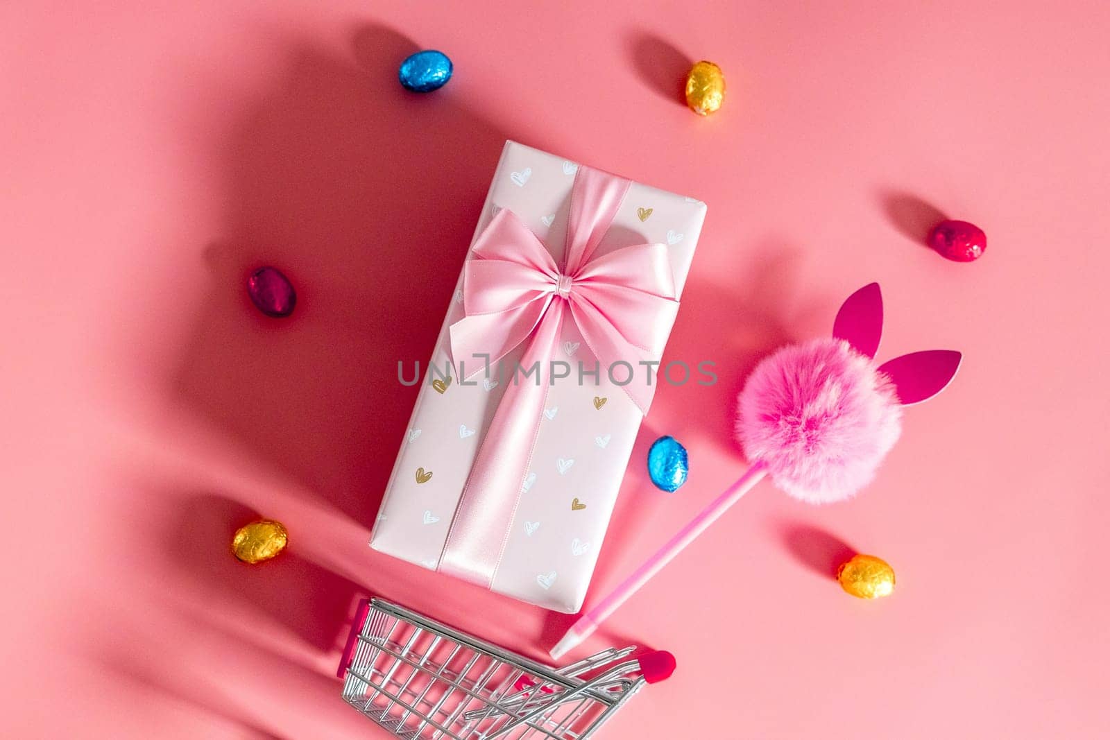 One large beautiful gift box with a bow, a fluffy bunny handle and chocolate Easter eggs in multi-colored wrappers fall out of a mini shopping cart on a pink background, flat lay close-up.