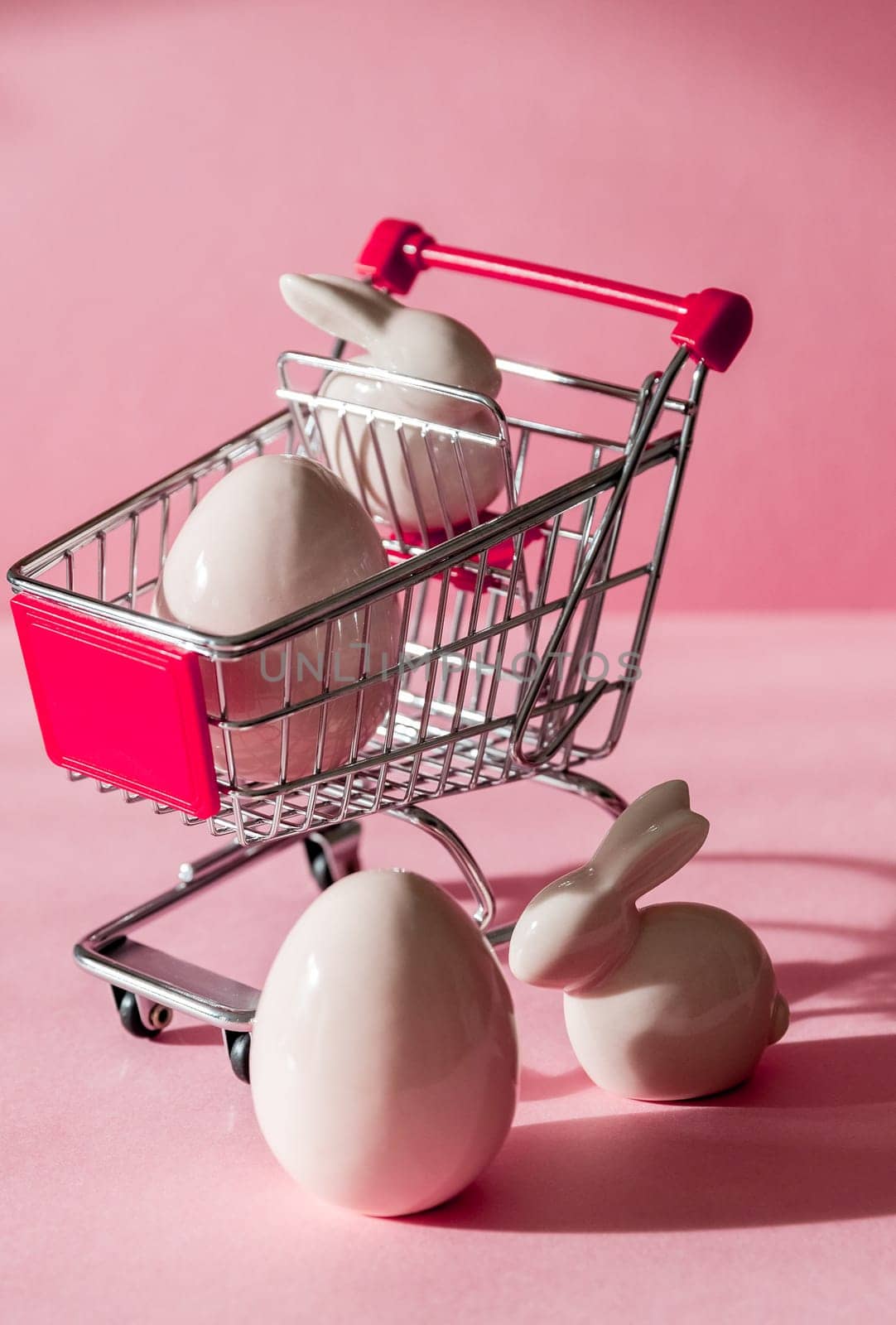 Porcelain figurines of Easter eggs and bunny with a mini shopping cart on a pink background, close-up side view.