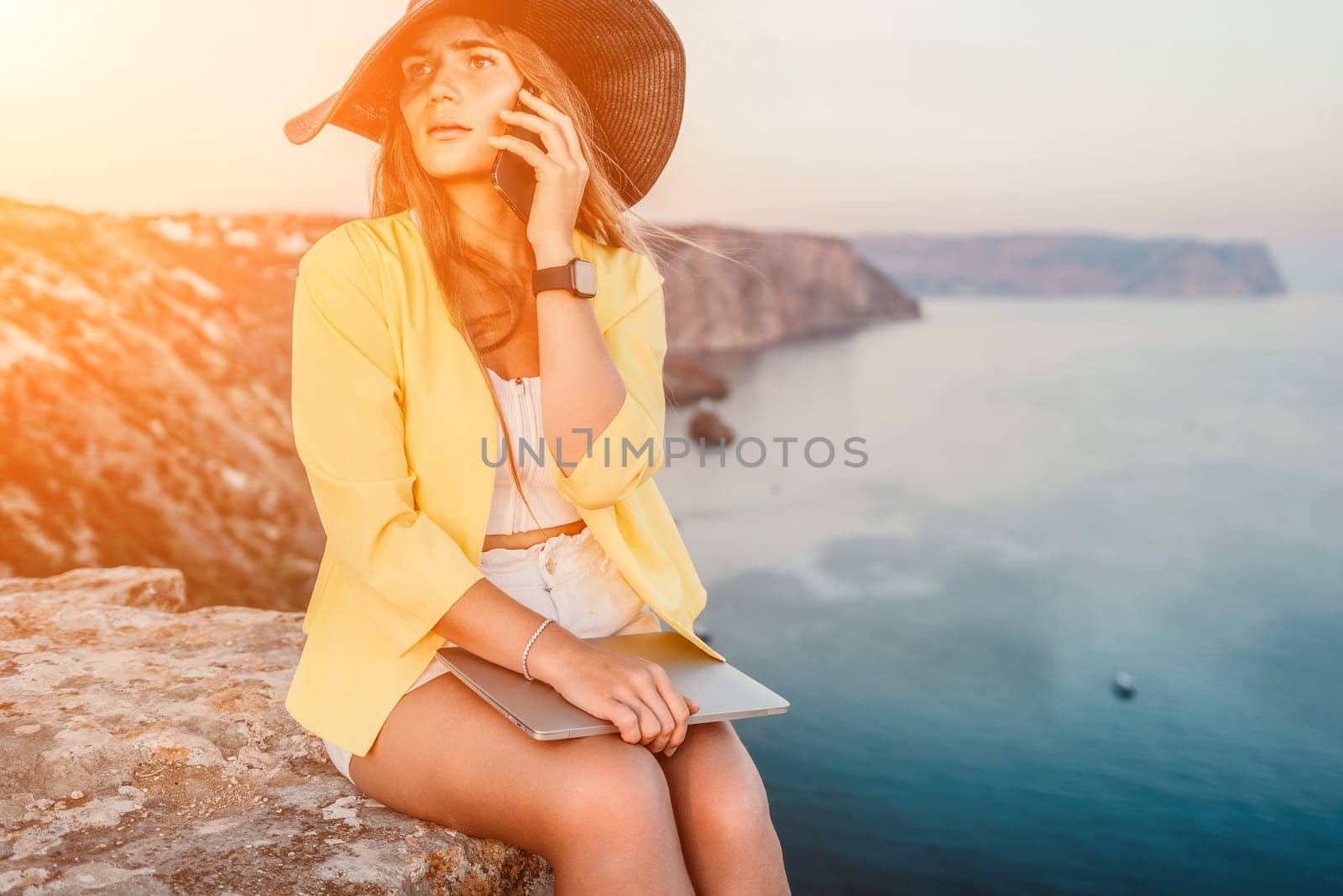 Successful business woman in yellow hat working on laptop by the sea. Pretty lady typing on computer at summer day outdoors. Freelance, travel and holidays concept.