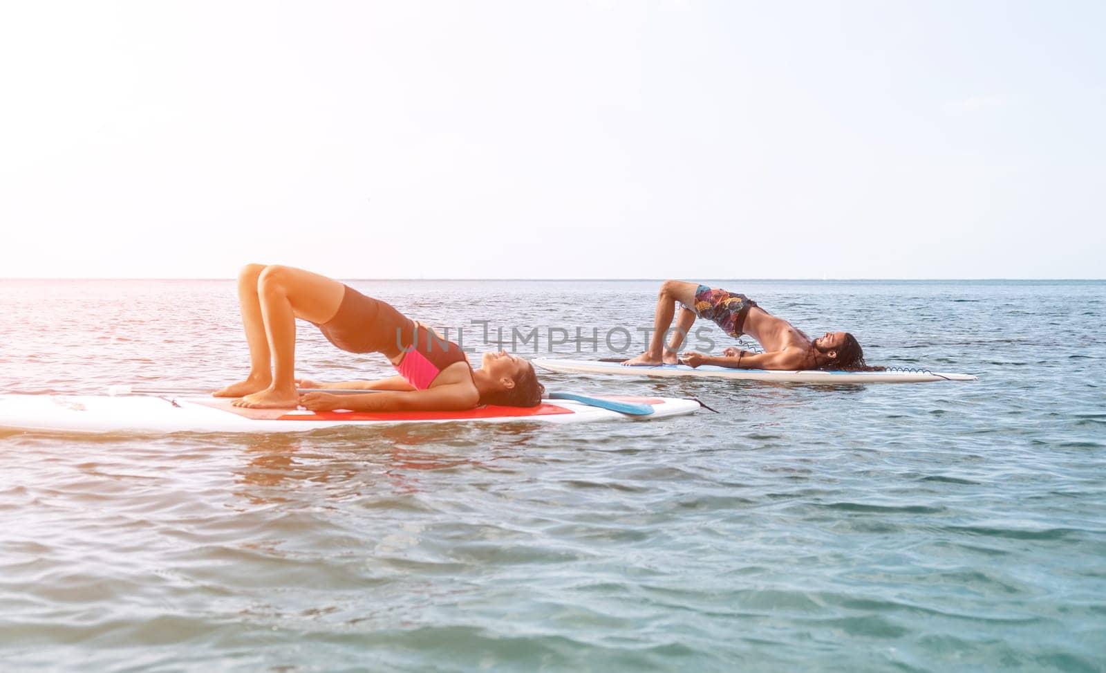 Woman man sup yoga. Happy young sporty couple practising yoga pilates on paddle sup surfboard. Female stretching doing workout on sea water. Modern family outdoor summer sport activity. by panophotograph