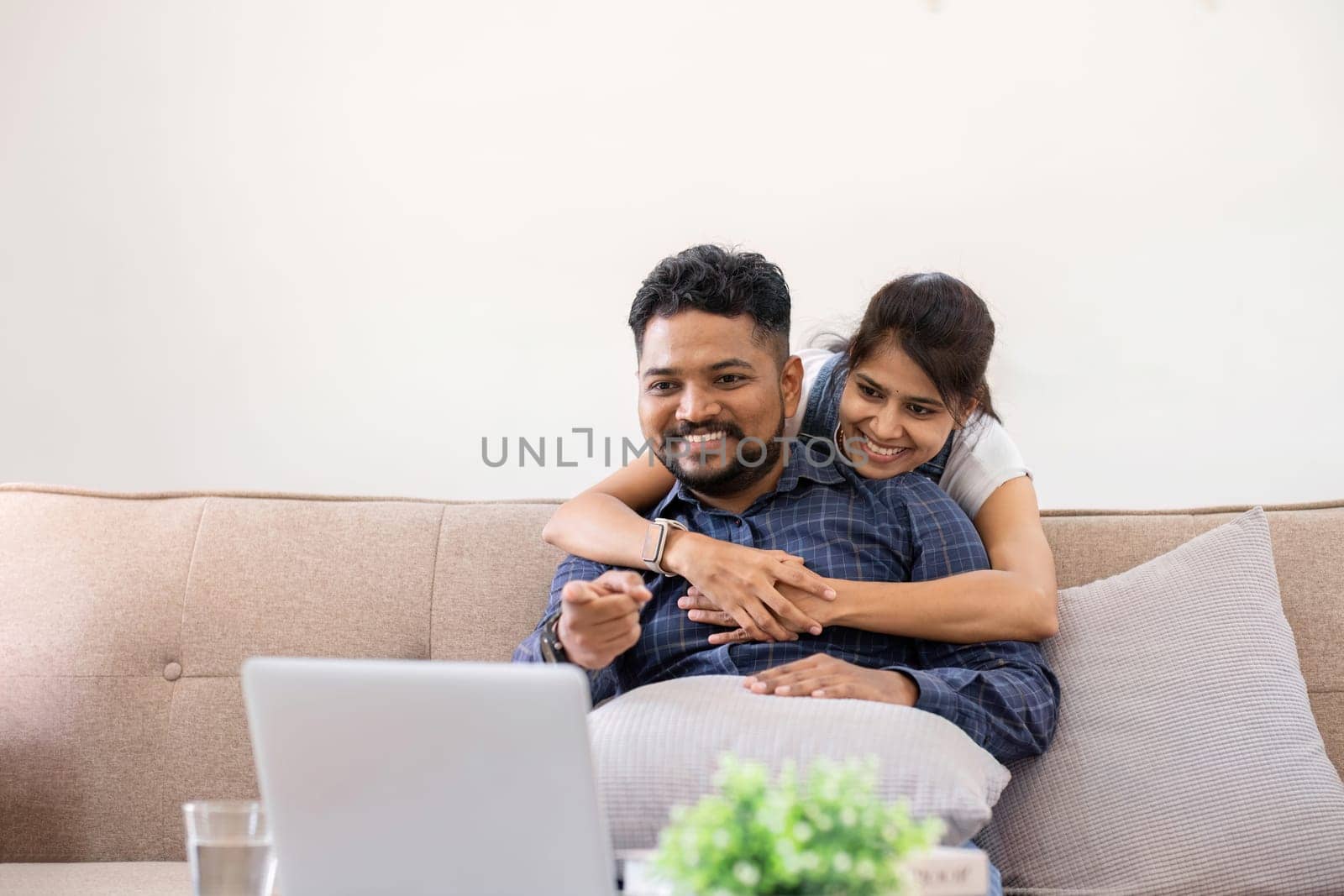 Cropped view of young woman hugging husband while using laptop on sofa in living room.