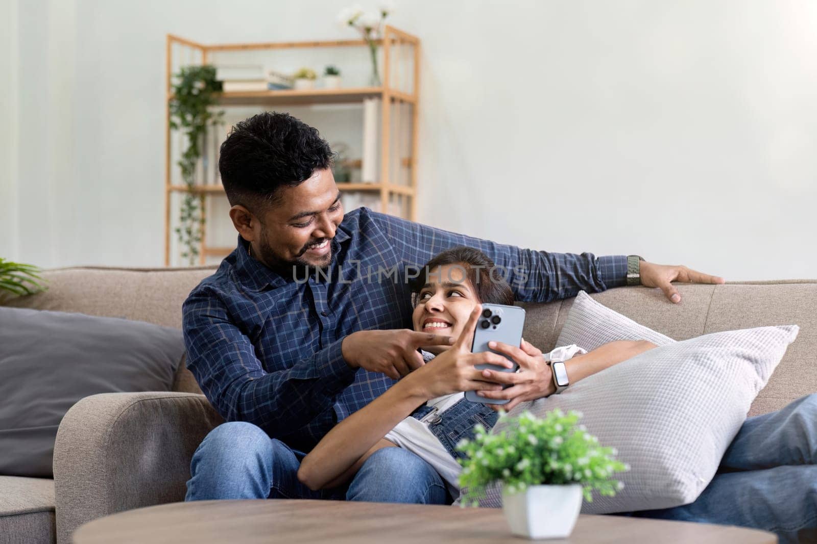 a couple sitting on the sofa looking at their mobile phones smiling and laughing together during happy holiday time.