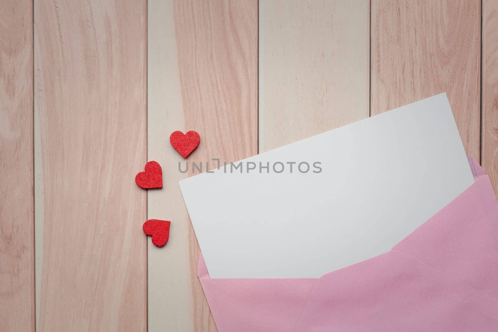 A pink envelope with a white piece of paper peeking out of the top with red heart-shaped confetti pieces on a light wooden table for love and Valentine's day concept