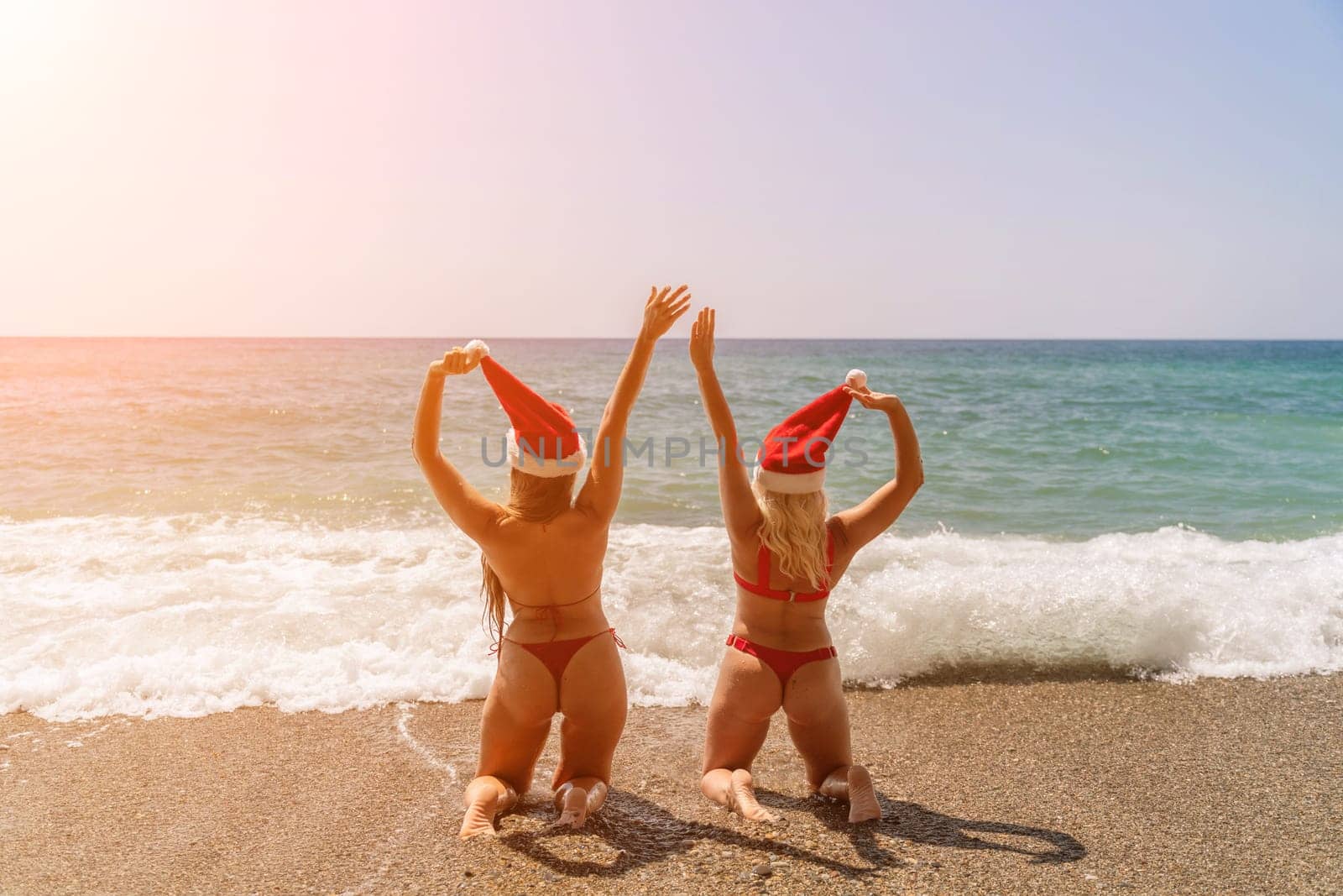 Women Santa hats ocean play. Seaside, beach daytime, enjoying beach fun. Two women in red swimsuits and Santa hats are enjoying themselves in the ocean, kneeling in the waves and raising their hands up. by Matiunina
