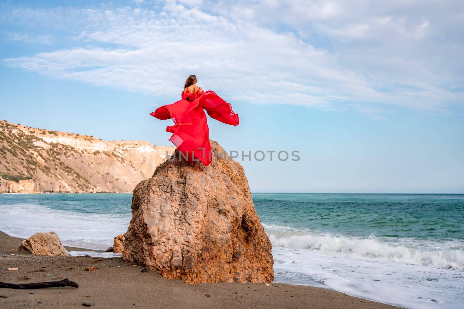 woman sea red dress. Woman with long hair on a sunny seashore in a red flowing dress, back view, silk fabric waving in the wind. Against the backdrop of the blue sky and mountains on the seashore