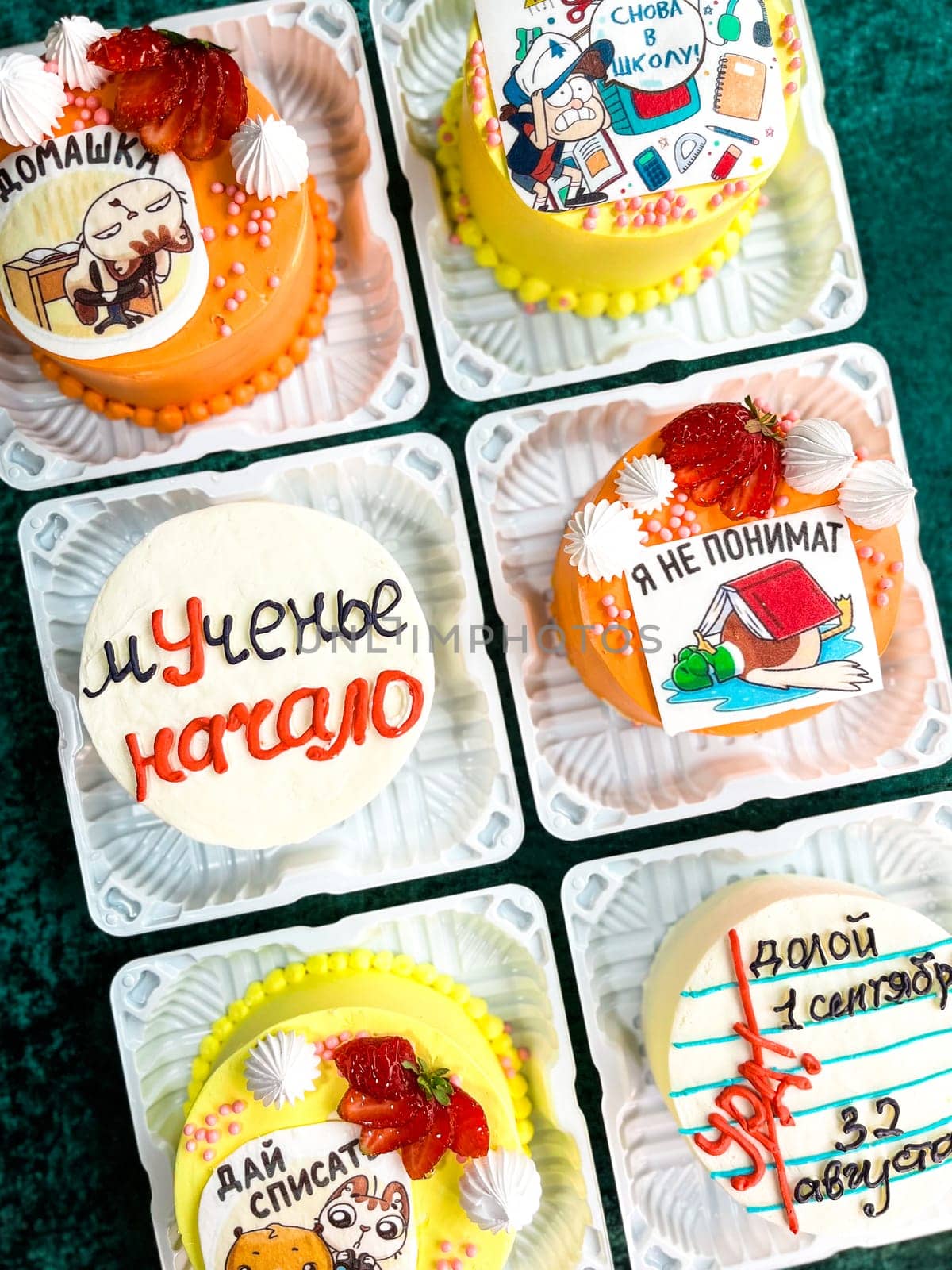 An assortment of six small square cakes with unique icing designs and decorations in plastic containers on a green surface, featuring cartoon characters, strawberries, and other decorative elements.