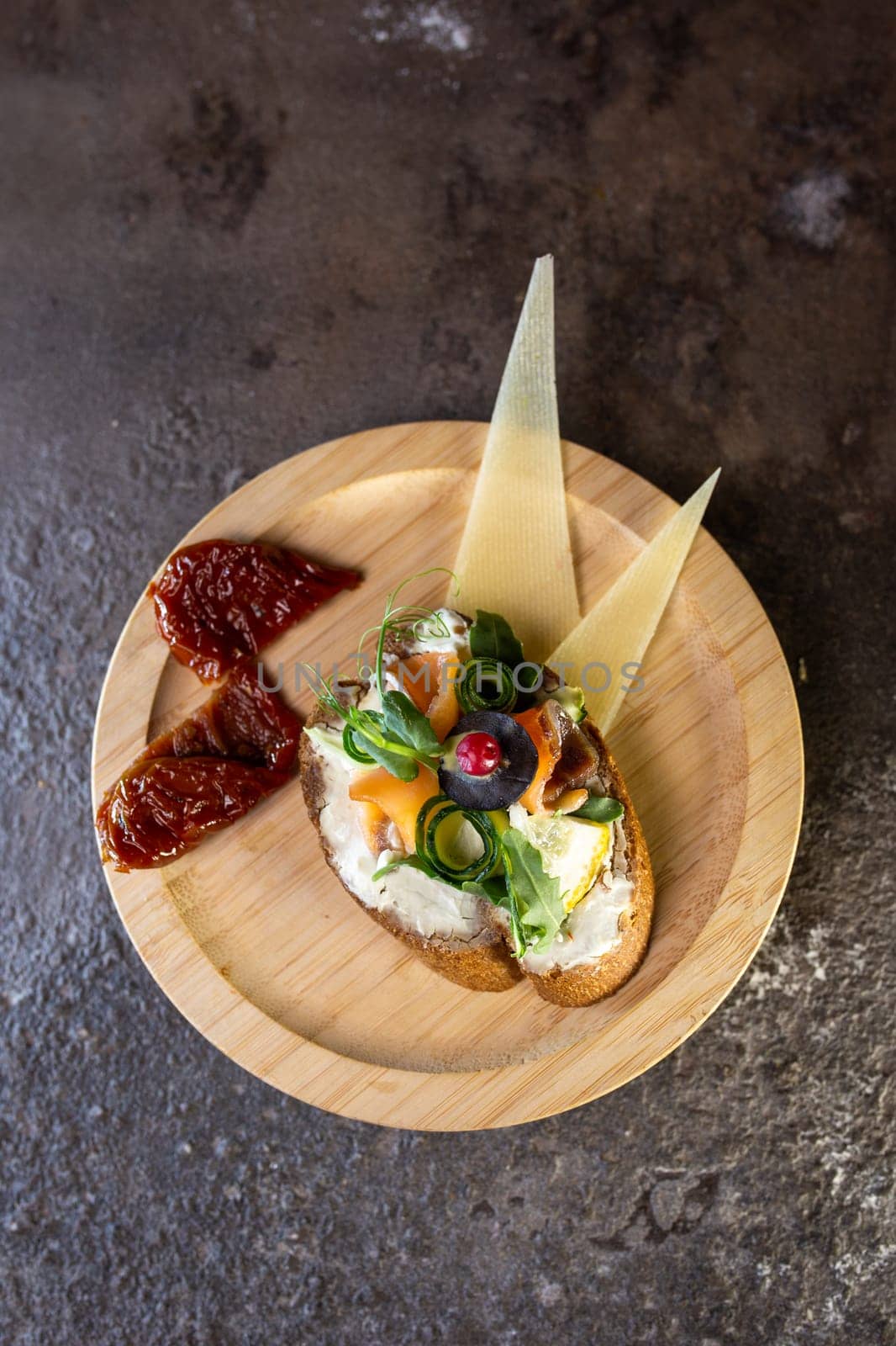 A delicious-looking open-faced sandwich made with cream cheese, cucumber, capers, and herbs on a wooden plate with cheese and sauce on the side. Perfect for a light lunch or snack.